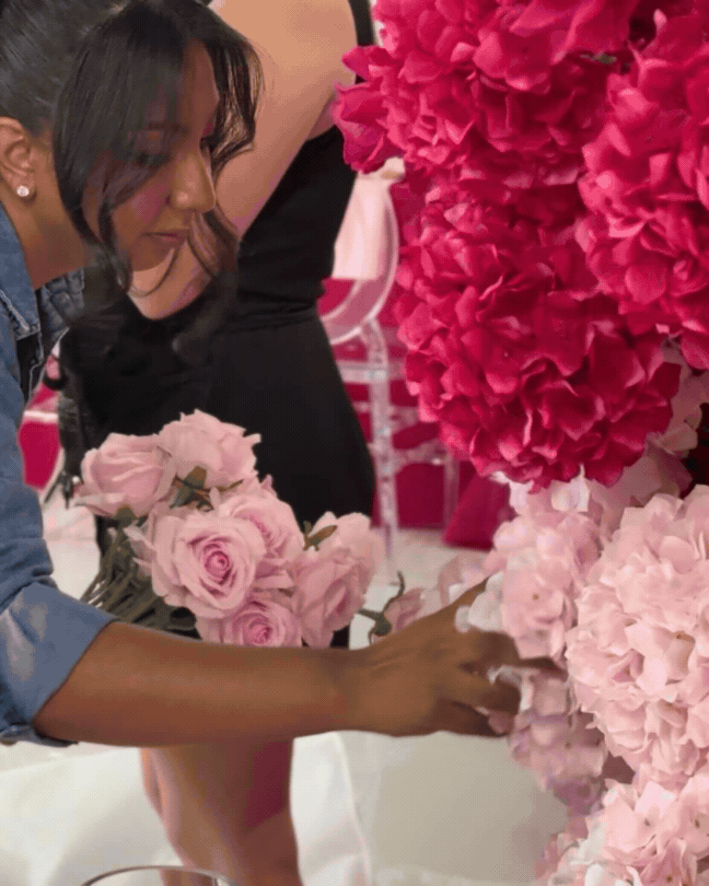 A woman with dark hair arranging pink and red flowers at a floral display, with chairs and decorations visible in the background.