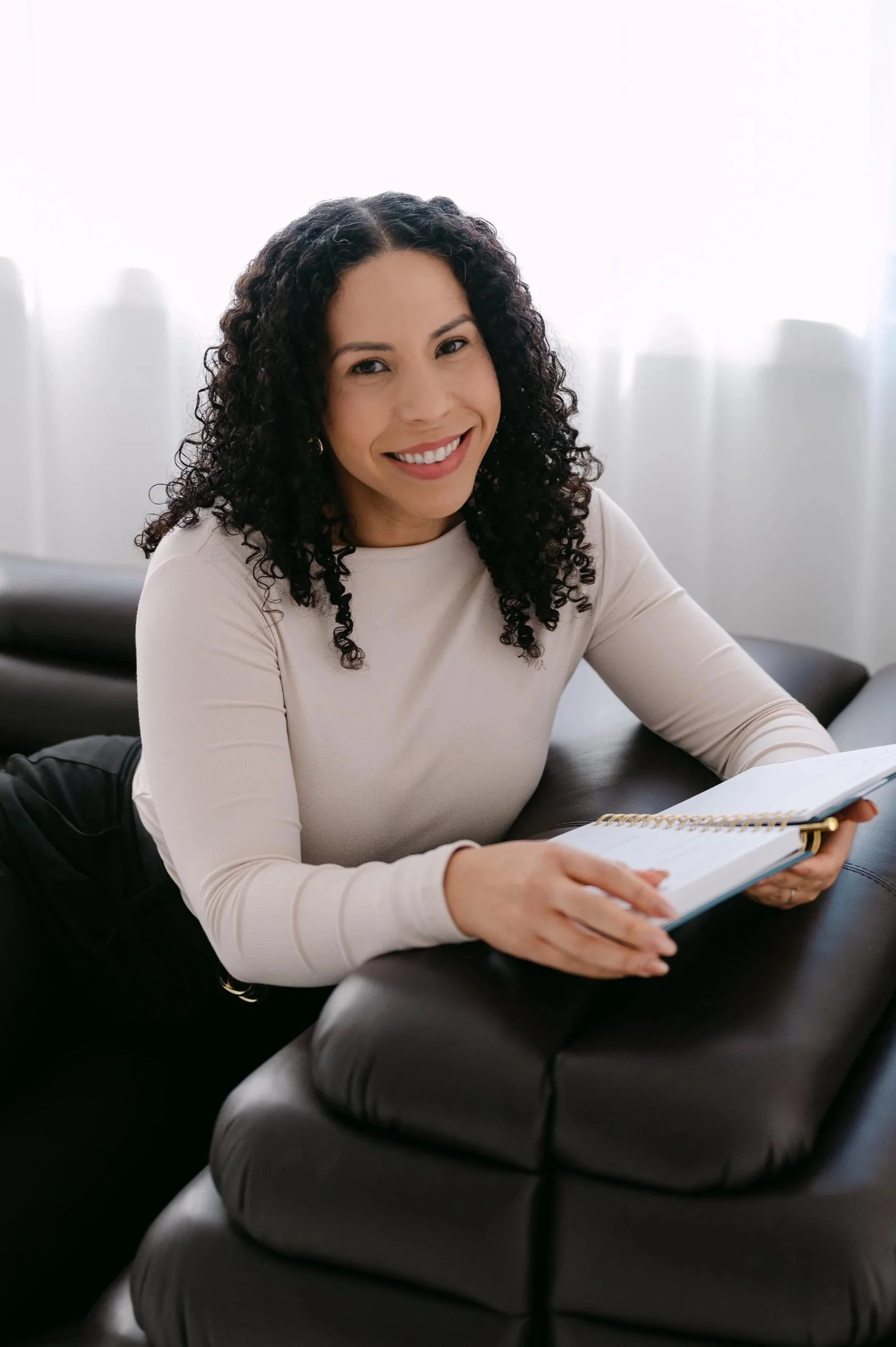 A woman with black curly hair sitting on a black leather sofa, smiling and holding an open notebook. She is wearing a light-colored long-sleeve top and has a bright, cheerful expression. The background features a white window with sheer curtains.