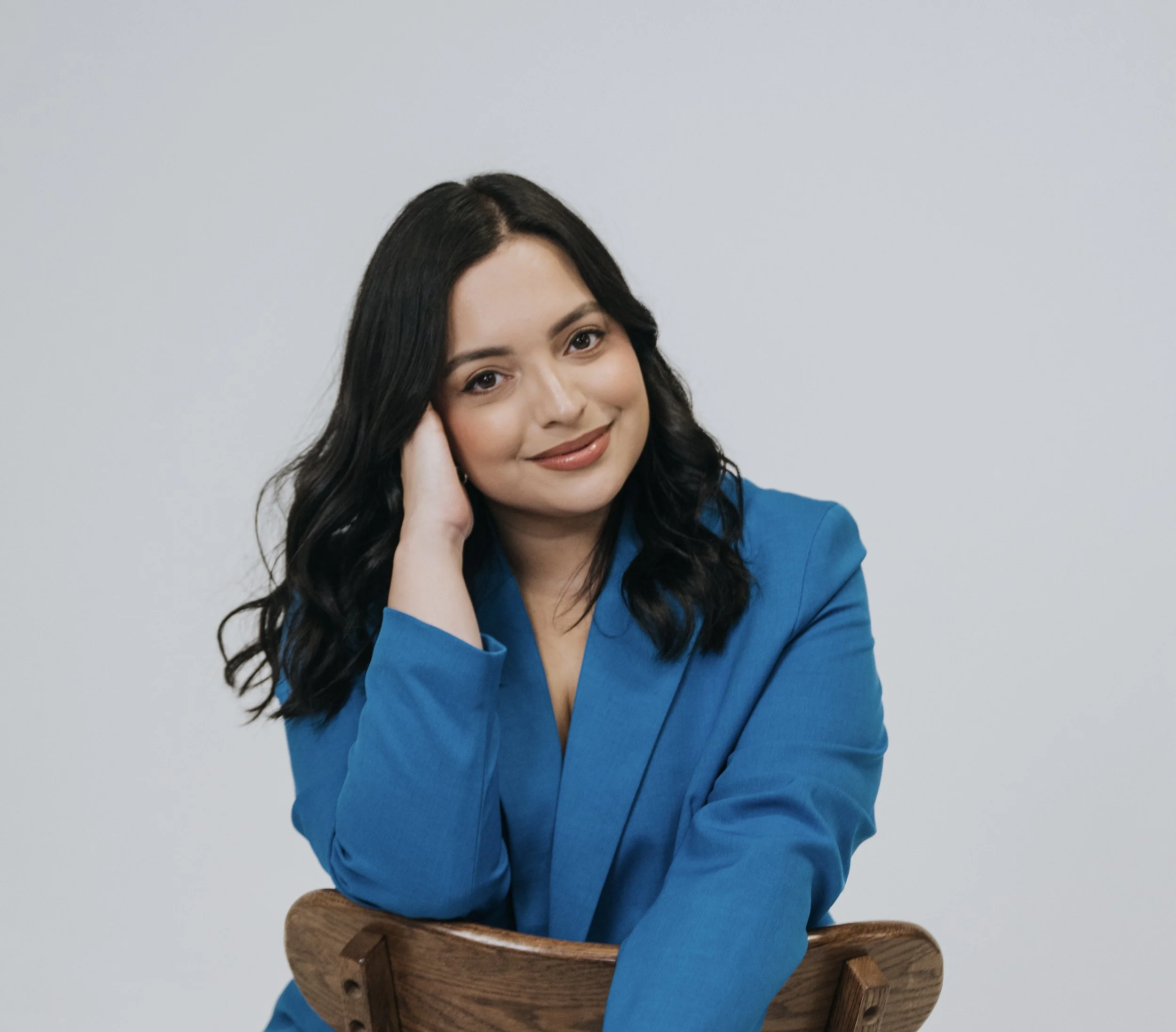 A woman with long dark hair sitting on a wooden chair, smiling and resting her head on her hand, wearing a blue blazer against a gray background.