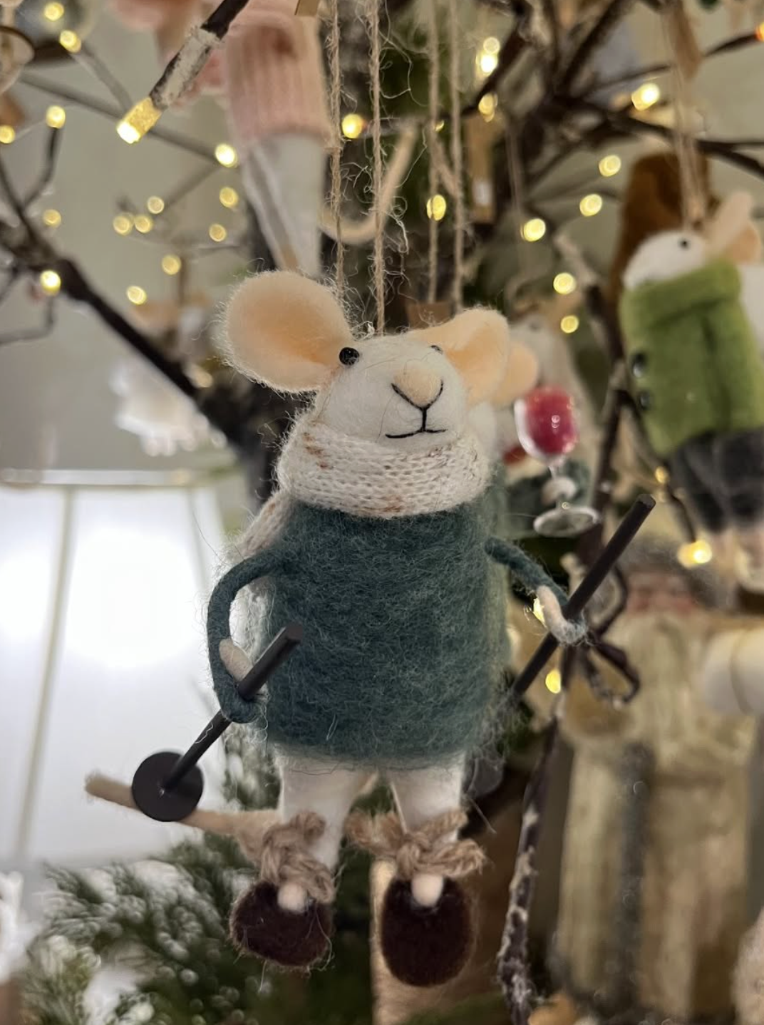 A cute mouse Christmas ornament wearing a green sweater and brown shoes, hanging from a decorated Christmas tree with lights.