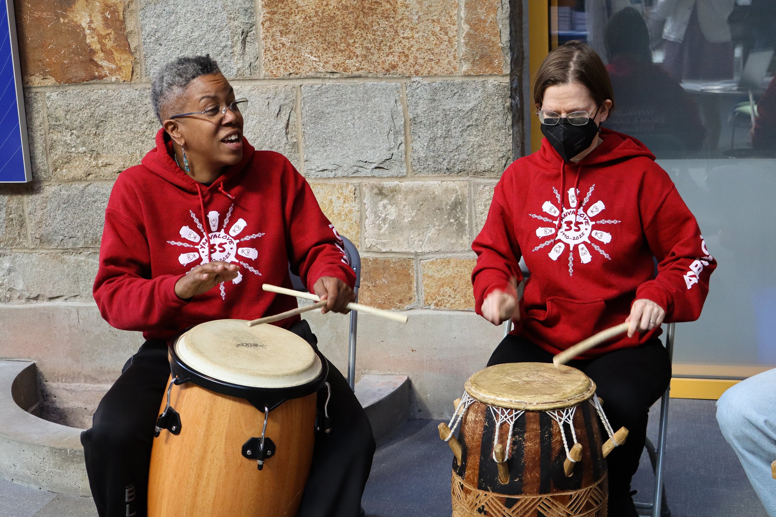 Two women wearing red hoodies with a white design are sitting and playing drums, one with a conga drum and the other with a djembe, against a stone wall background.