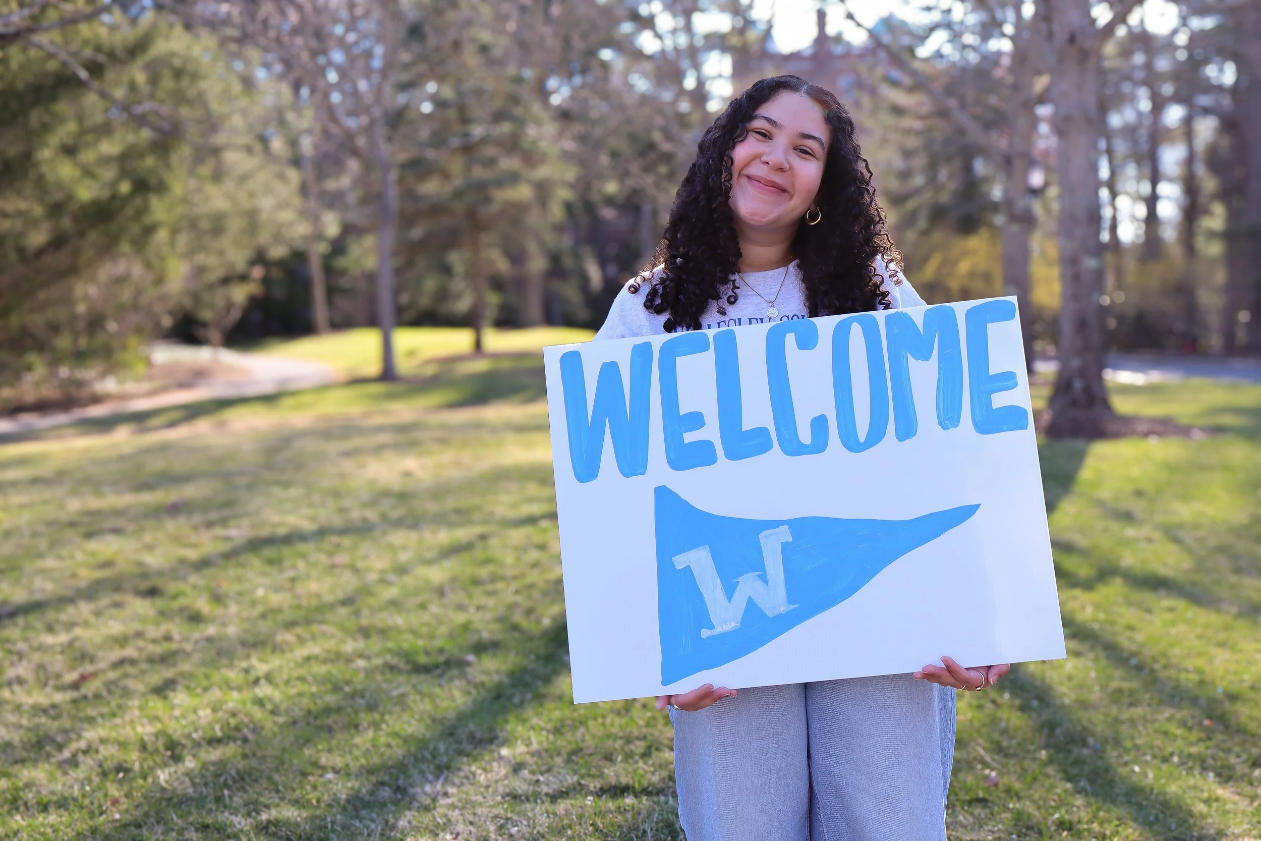 A young woman with curly dark hair smiling and holding a large white sign with blue writing that says "WELCOME" in a park with trees and grass in the background.