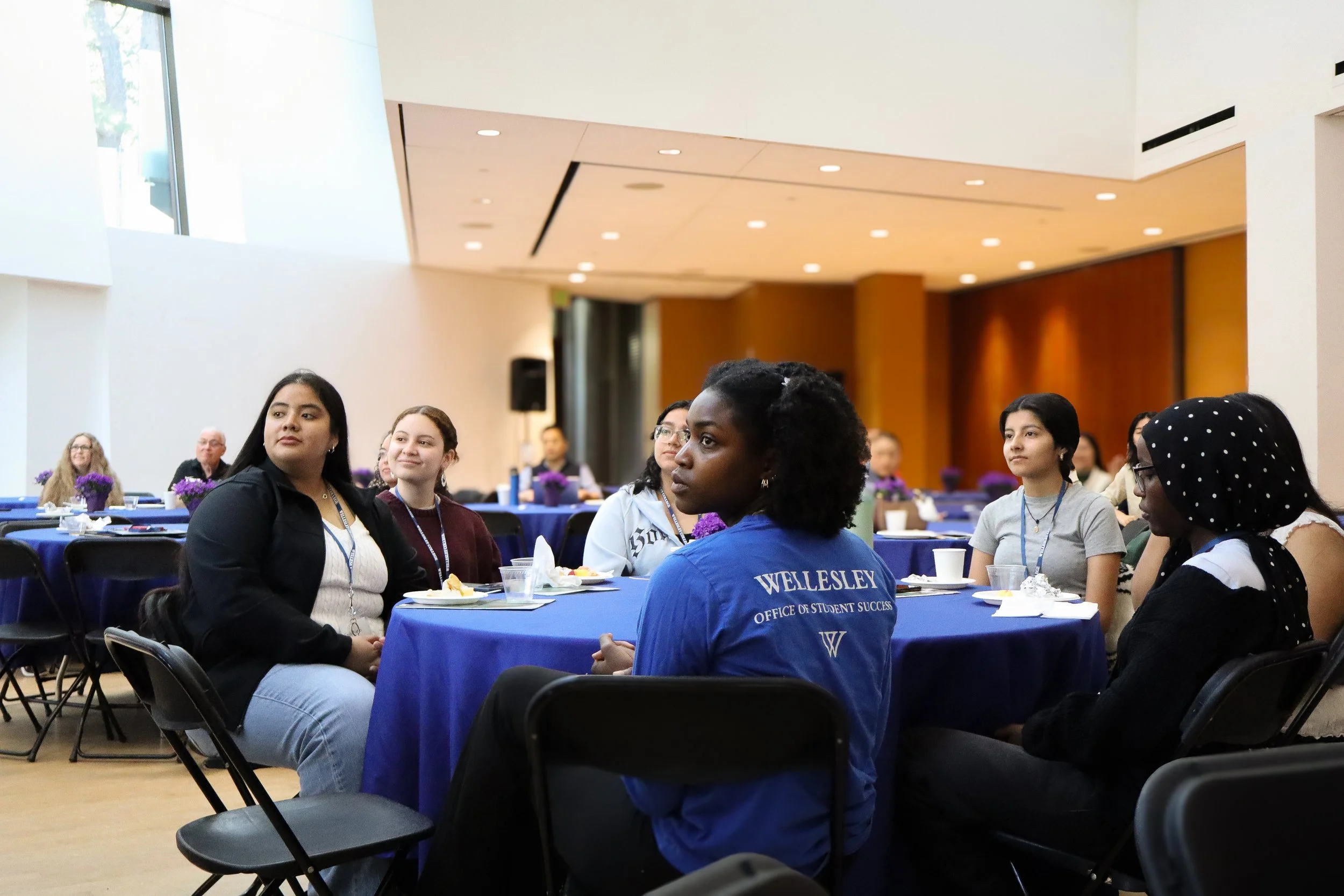 Group of diverse young women and a few men seated at round tables with blue tablecloths, listening attentively in a large, well-lit conference room with wooden panels, during a formal event or workshop.