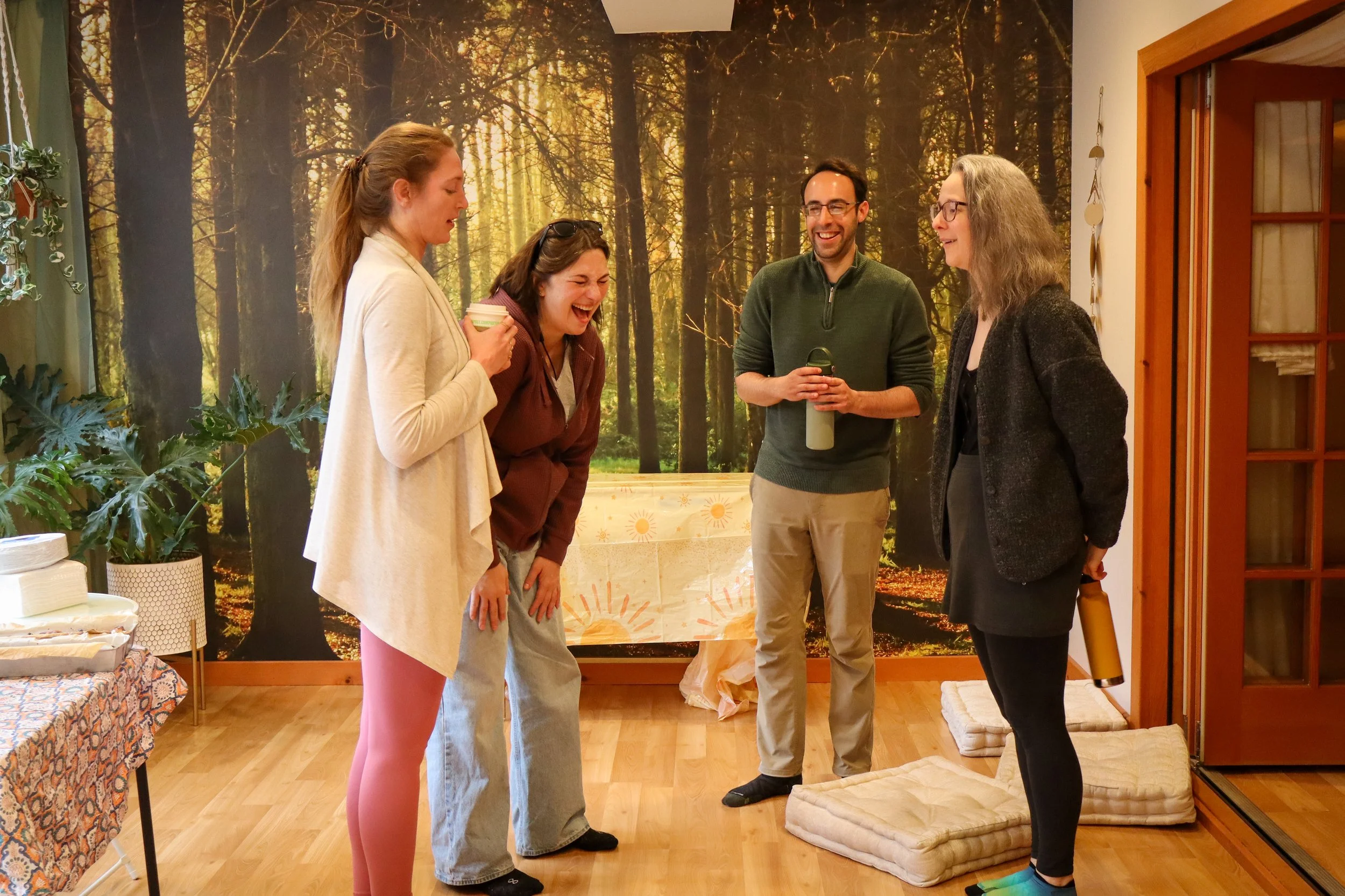 A group of five people laughing and talking indoors with a forest-themed mural in the background. They are standing on a wooden floor, with some cushions and tablecloths nearby.