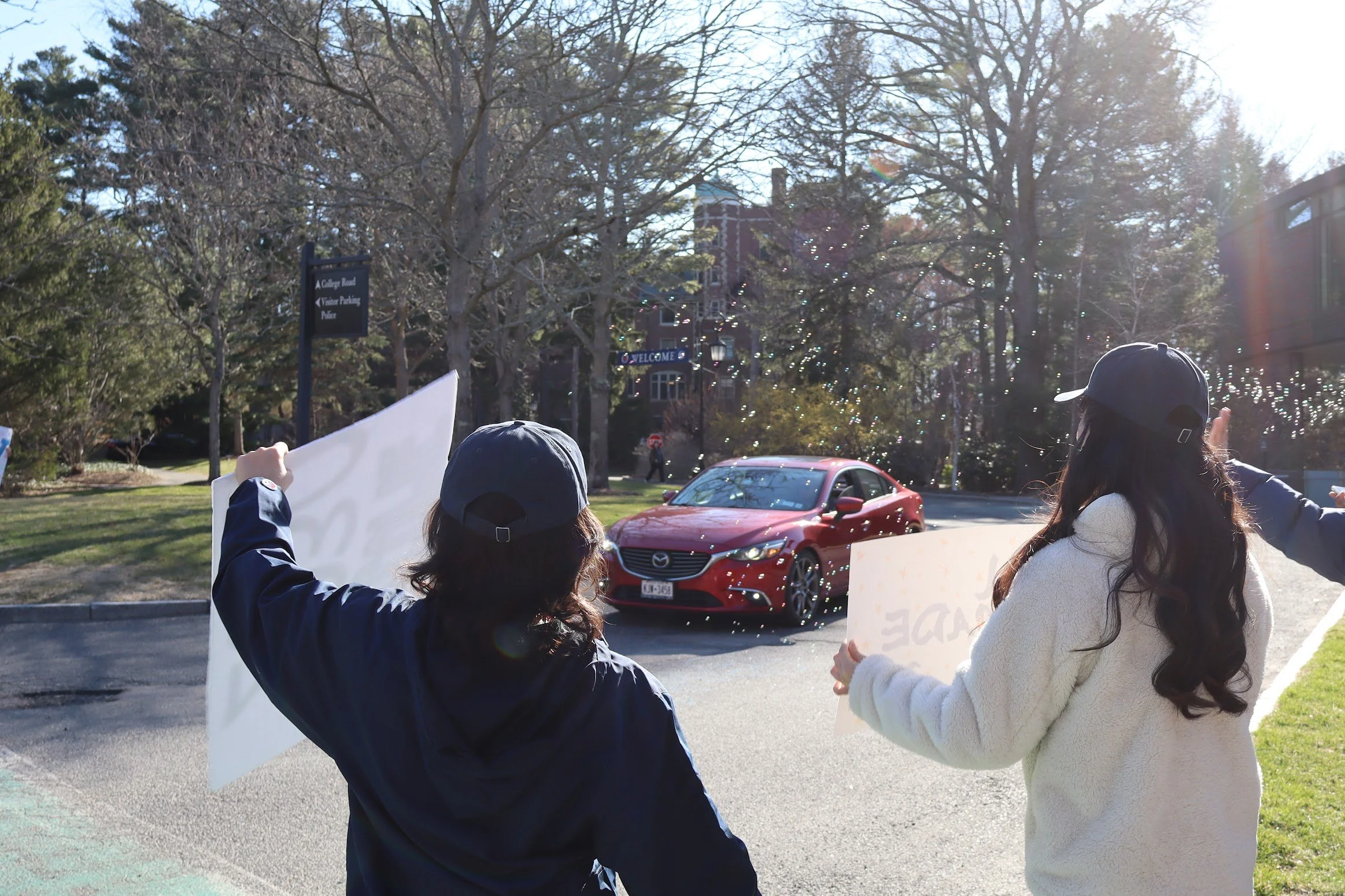 People participating in a protest or demonstration outdoors, holding signs with one person wearing a black jacket and another in a white sweater. A red Mazda car is parked on the street with trees and buildings in the background.