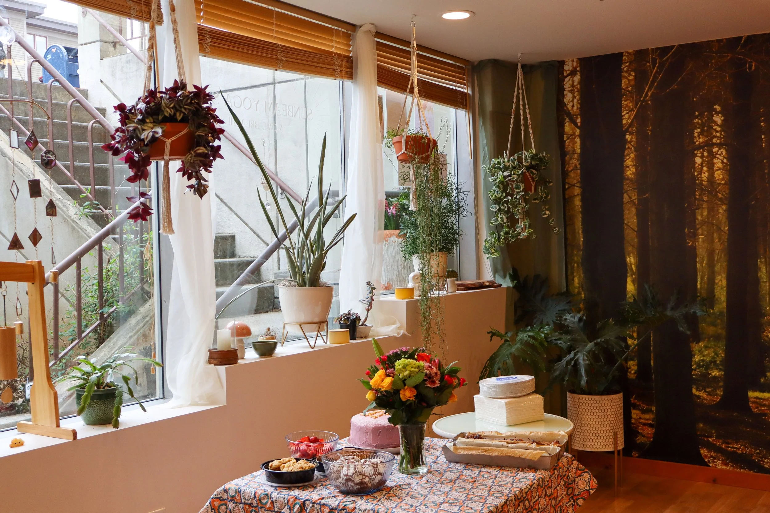 Inside a cozy room with a large window, decorated with plants hanging and placed on the windowsill. There is a table with a colorful patterned tablecloth, holding a bouquet of flowers, bowls of snacks, and a pink cake. The backyard and autumnal fores