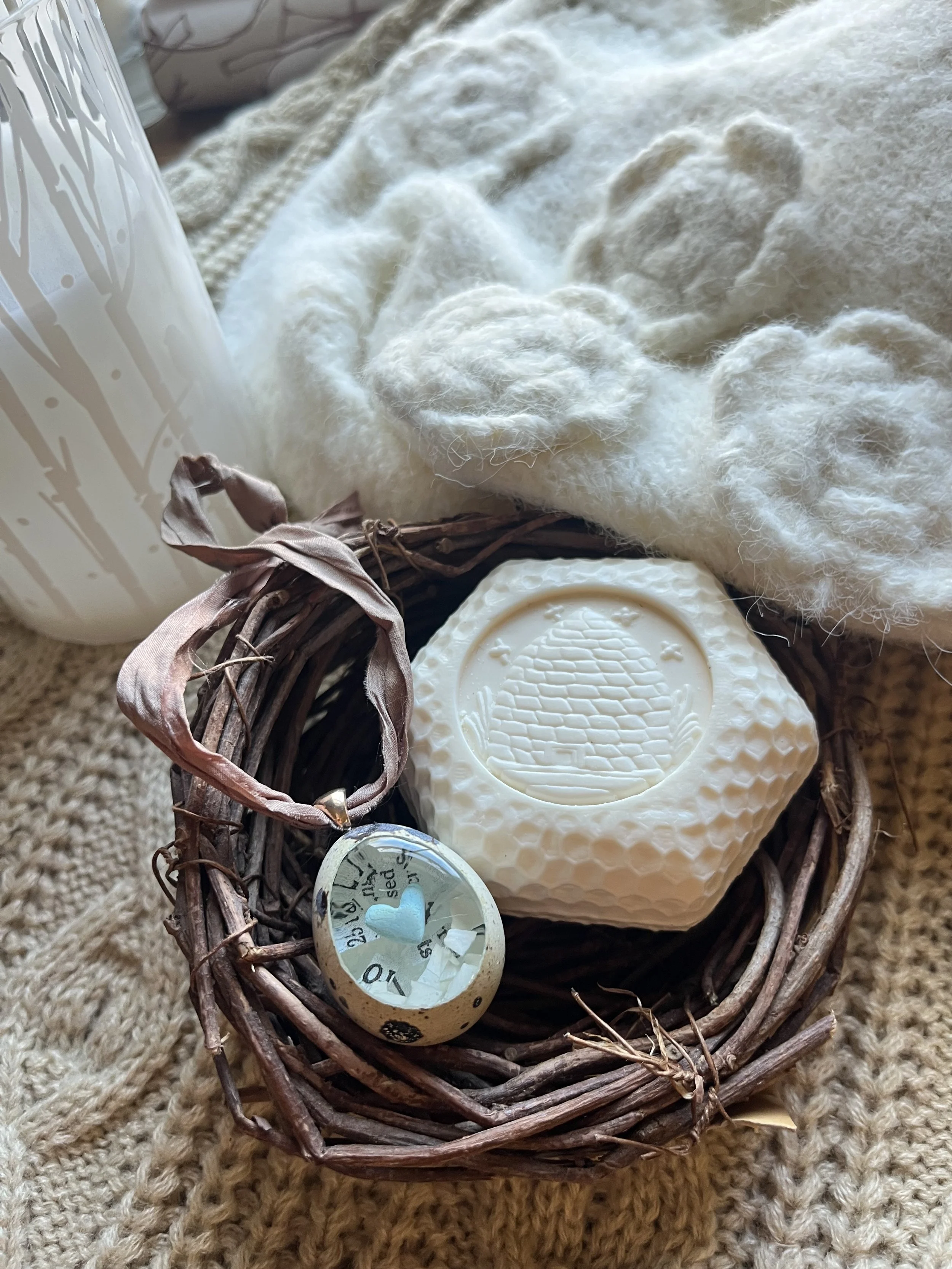 A small woven basket contains a cream-colored decorative soap with a textured circular pattern on top, a white ceramic candle with a leaf pattern, a small grey ceramic heart, and a white circular ornament with black markings and text, all placed on a beige knitted surface.