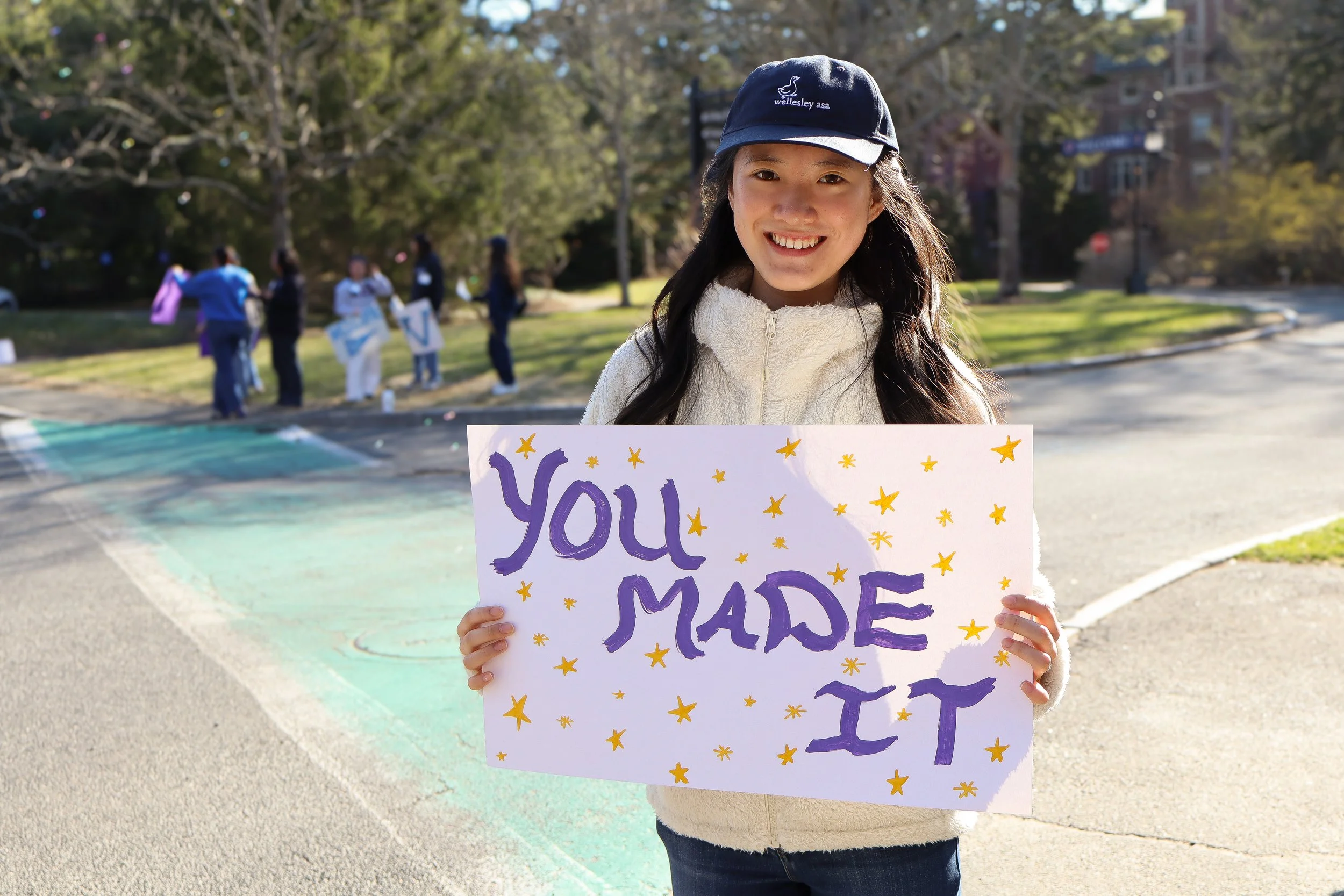 A young girl smiling outdoors, holding a sign that says 'You Made It' with yellow star decorations, wearing a navy hat and a cream fuzzy jacket.