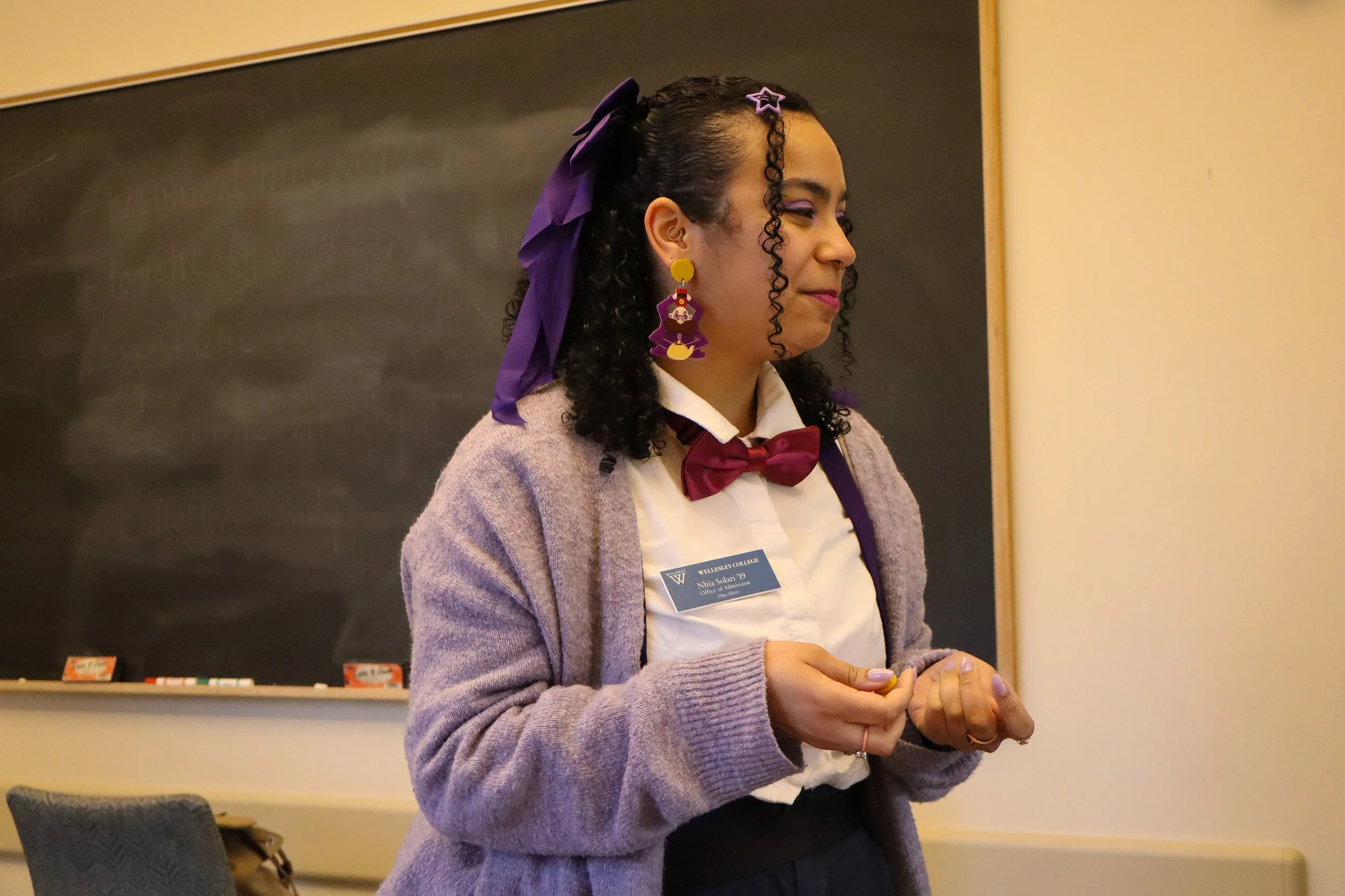 A woman with curly dark hair wearing a purple bow and star-shaped earrings, dressed in a beige cardigan and a white shirt with a red bow tie, standing in front of a blackboard in a classroom or lecture hall.