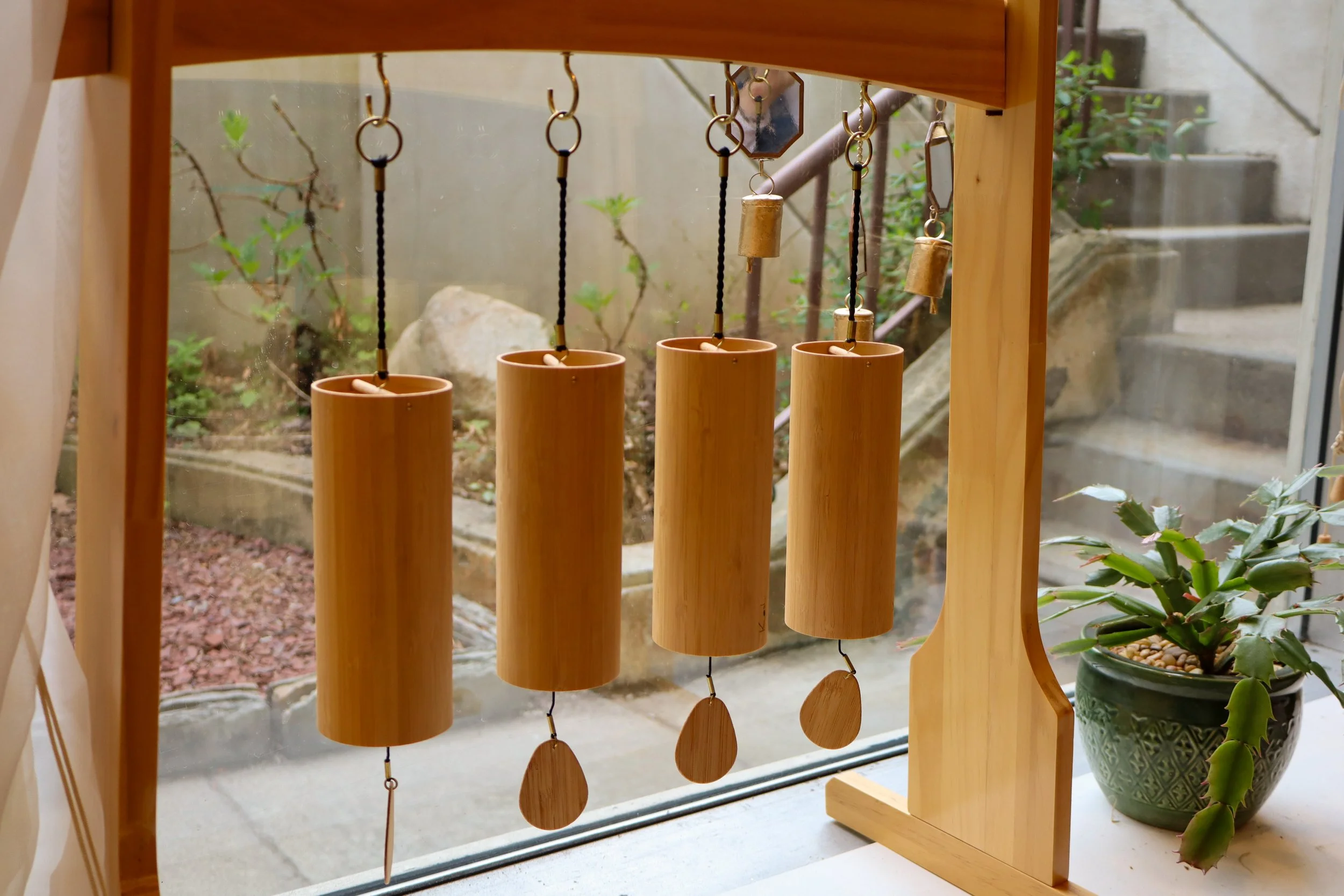 A wooden wind chime with five bamboo tubes hanging inside a window, with a garden and concrete stairs outside.