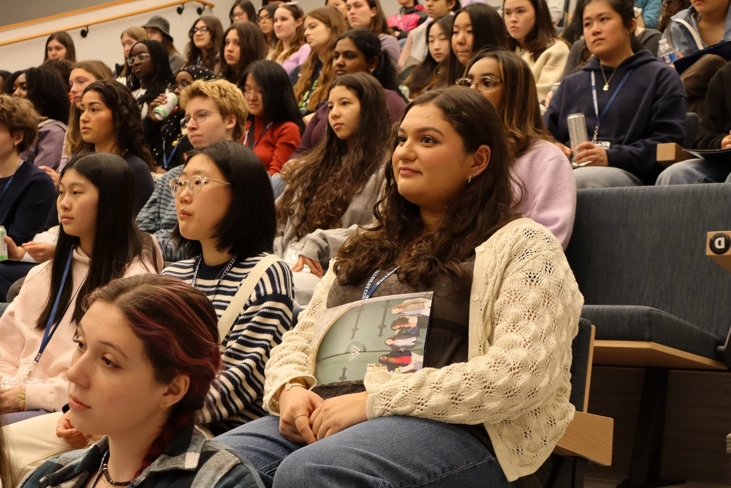 A diverse group of students sitting in an auditorium, attentively listening during a presentation or lecture, some holding notebooks or water bottles.