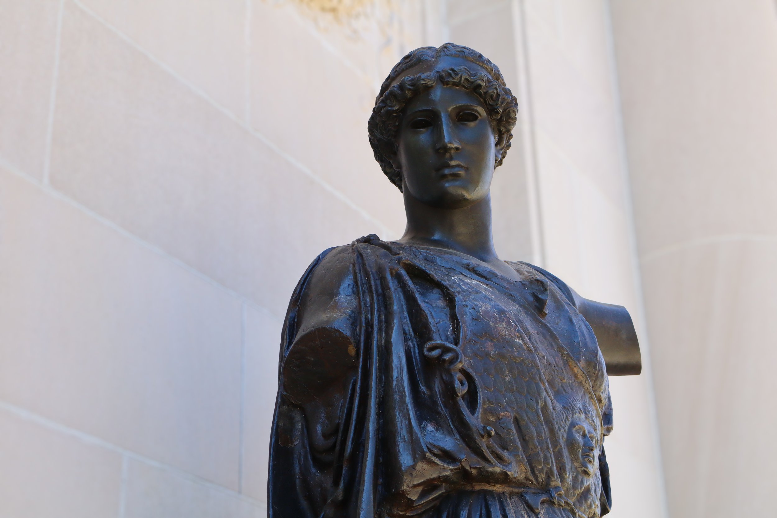 A bronze sculpture of a woman with curly hair, wearing a dress, standing against a light-colored tiled wall.