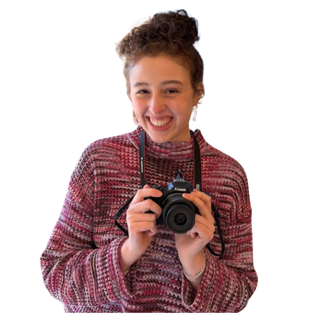 Young woman with curly hair smiling and holding a camera, wearing a multicolored sweater.