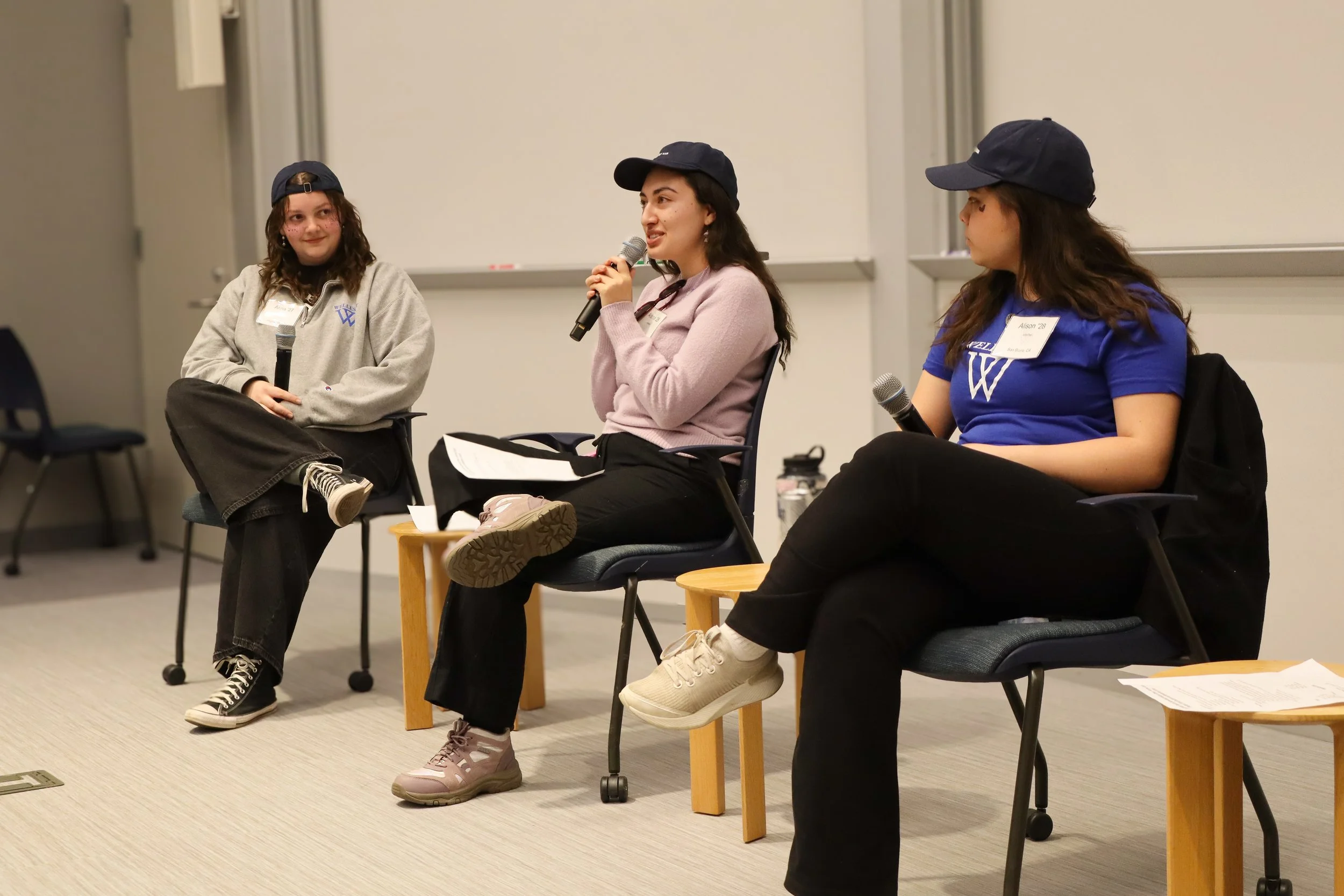 Three young women sitting on chairs in a panel discussion facing an audience, each holding a microphone, with a whiteboard behind them.