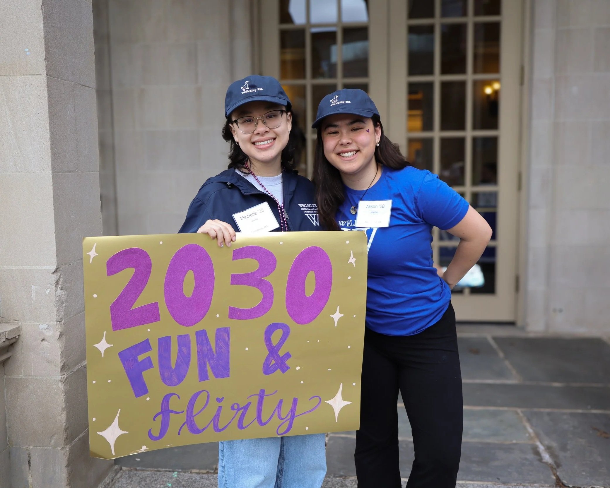Two young women smiling and standing outside, holding a colorful sign with purple and yellow text. They are wearing matching blue caps and name tags that indicate they are attending an event. The background shows a building with large windows and bei