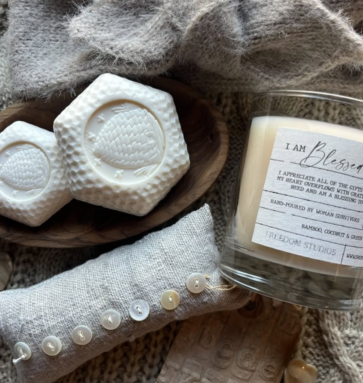 A cozy scene with two hexagonal soaps with embossed strawberry designs, a wooden bowl, a candle labeled 'I am blessed' with a gratitude message, a fabric-covered button strip, and a wooden stamp on a textured blanket.