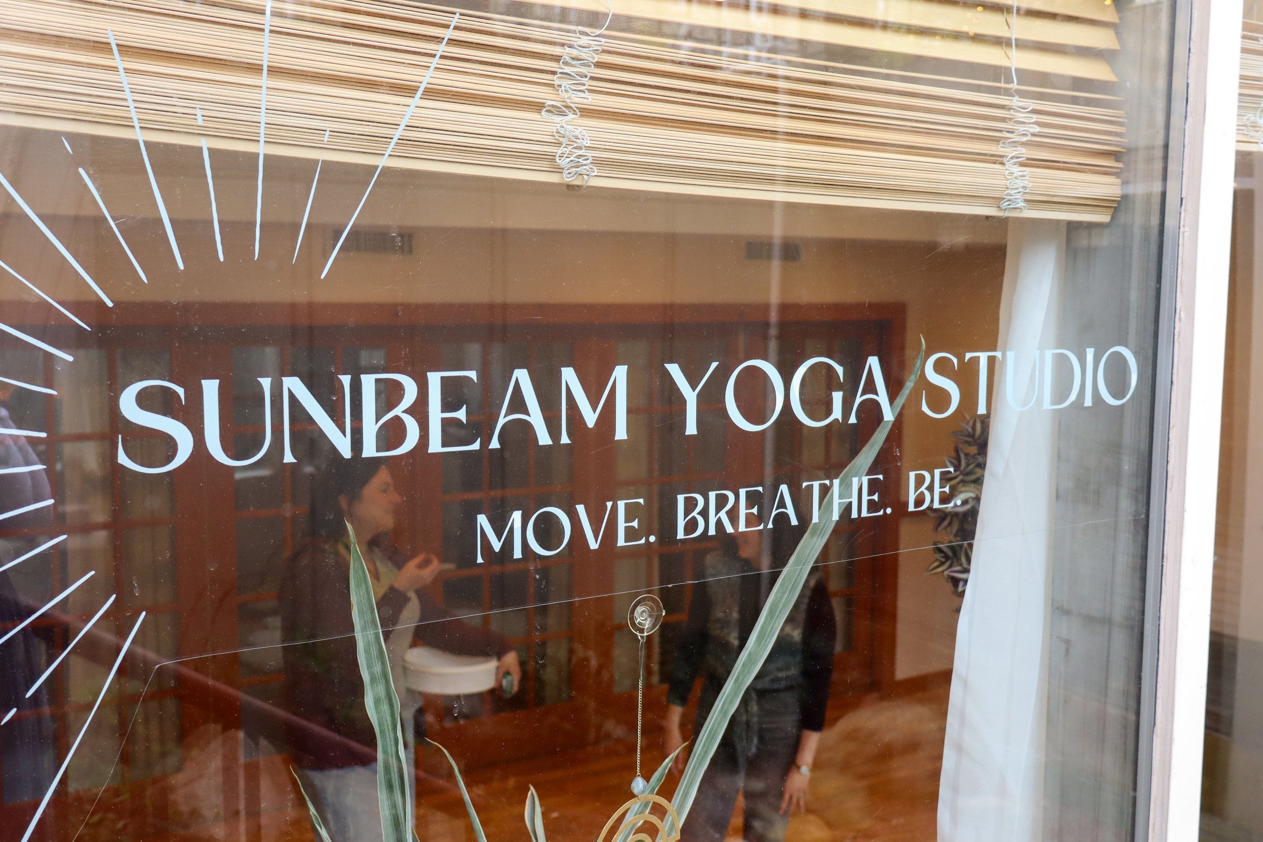 The window sign reads "SUNBEAM YOGA STUDIO" and has the partial words "MOVE. BREATHE. BE." beneath it. Reflections on the glass show several people inside the studio.