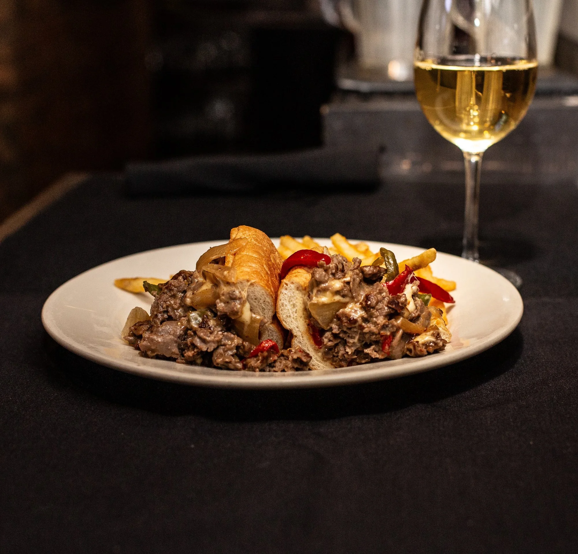 A plate with a beef-stuffed bread roll, French fries, and some red and green peppers. A glass of white wine is in the background on a black table.