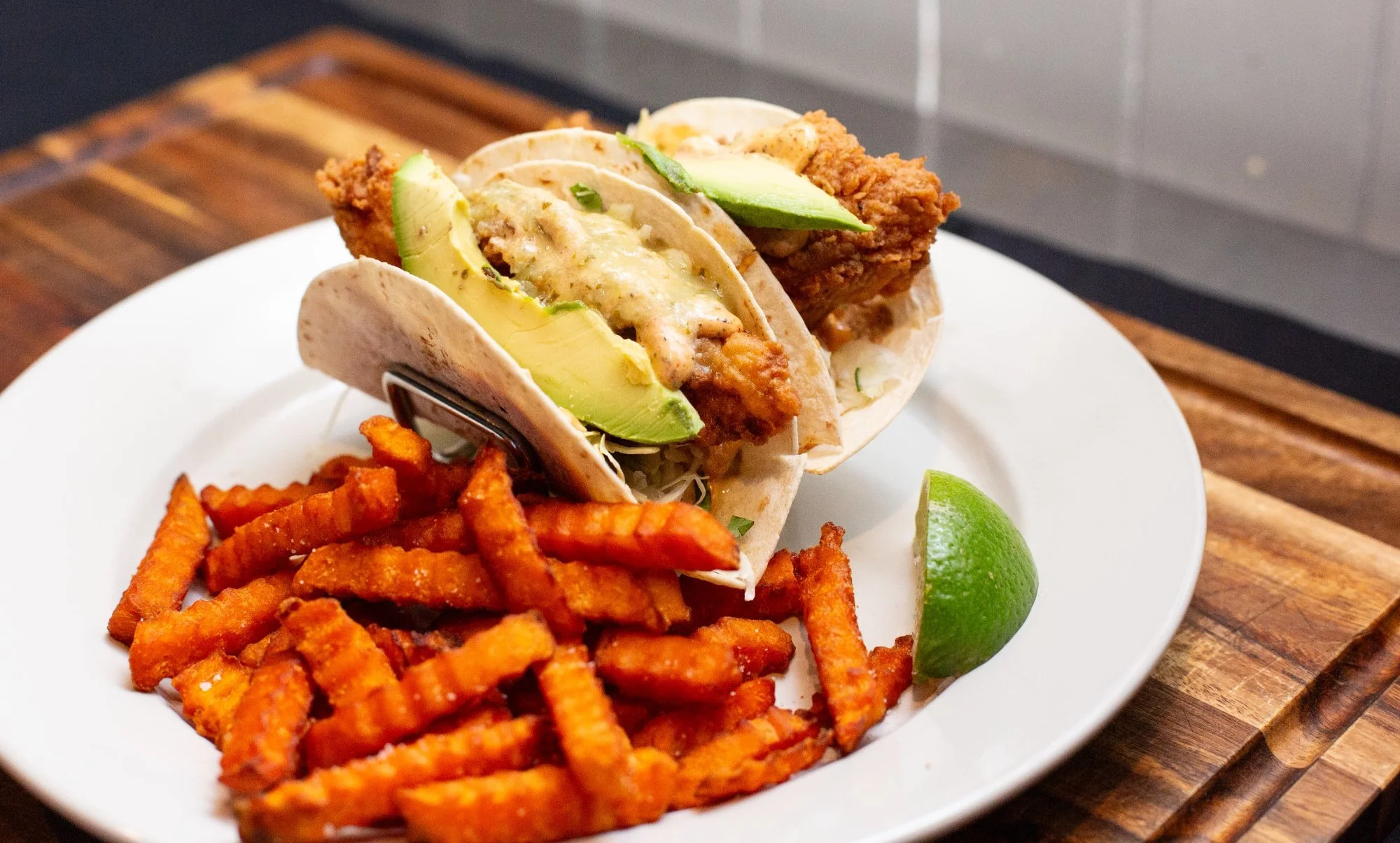 Tacos filled with fried chicken, avocado, and sauce, served with sweet potato fries on a white plate and a lime wedge.