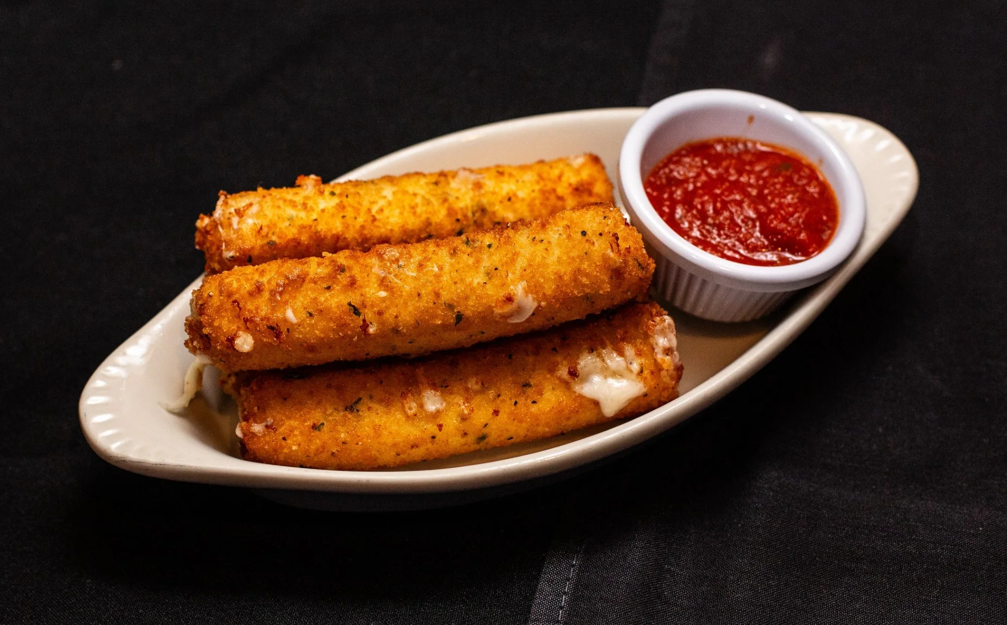 Three mozzarella sticks with a side of marinara sauce on a white oval dish against a black background.