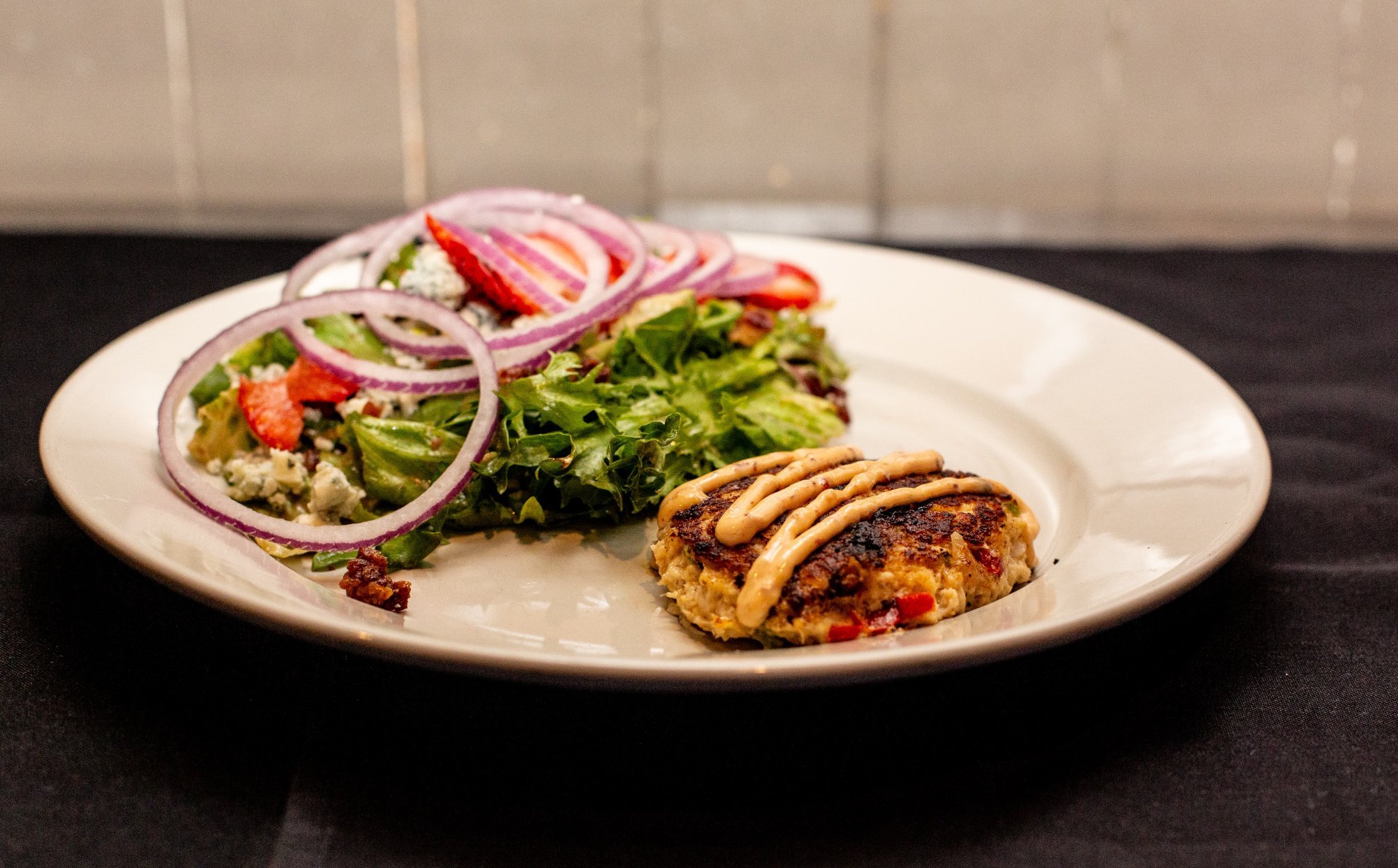 Crab cake plate with salad topped with red onion slices and a grilled patty with sauce, on a black table.