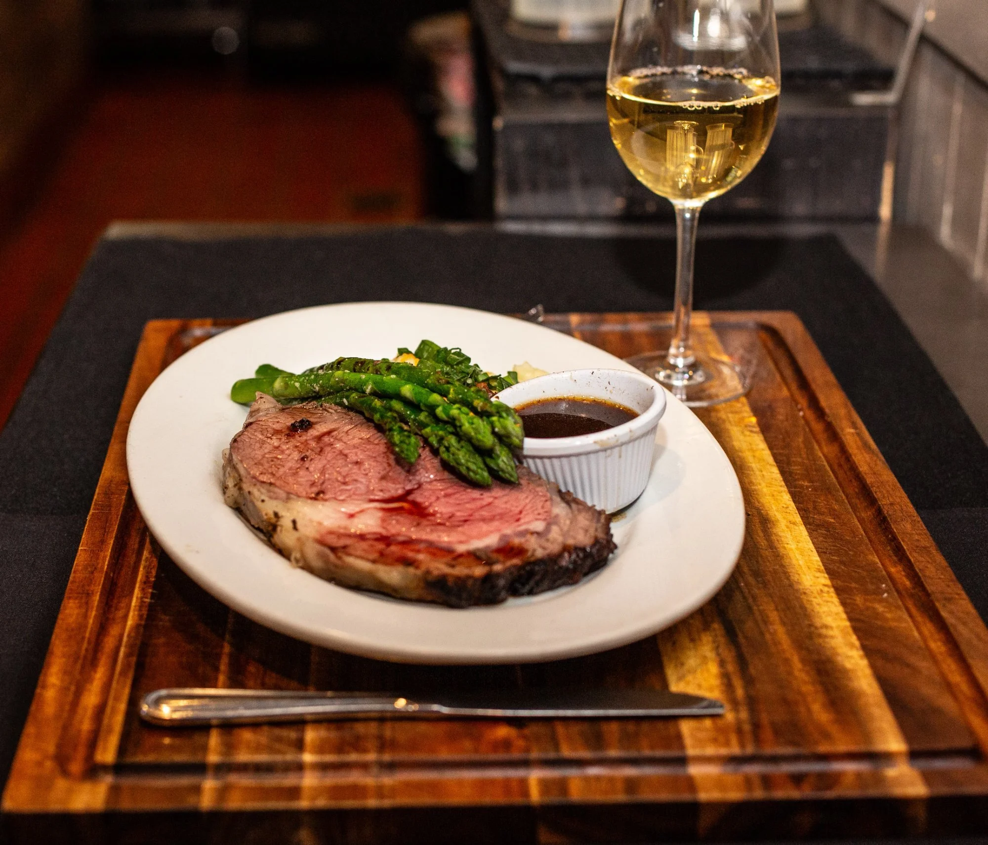 A plate with a cooked steak, asparagus, and a bowl of sauce, accompanied by a glass of white wine.