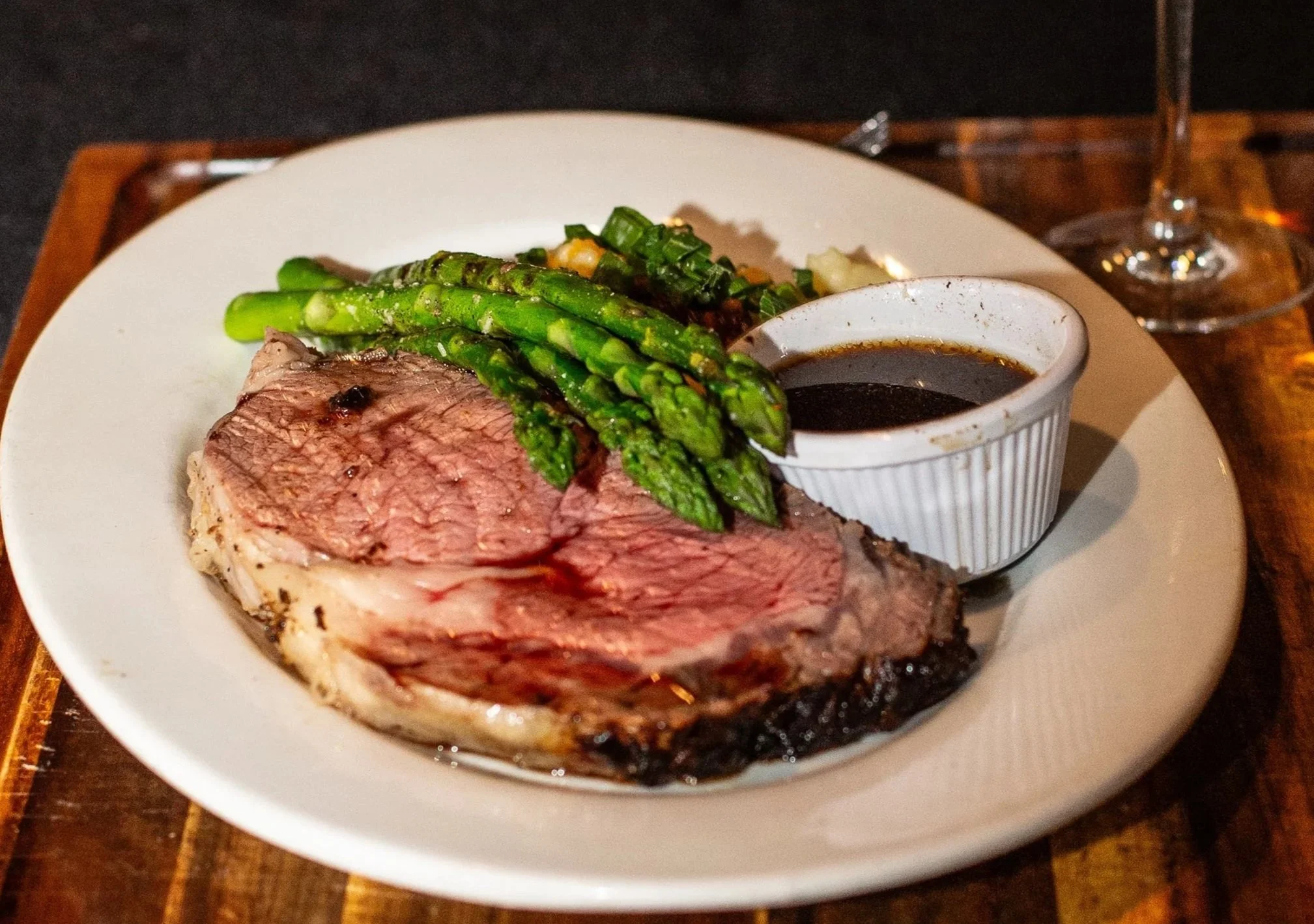 A plate of cooked Prime Rib with grilled asparagus, a small cup of dark gravy, and some chopped vegetables, served on a white plate with a clear cocktail glass in the background.