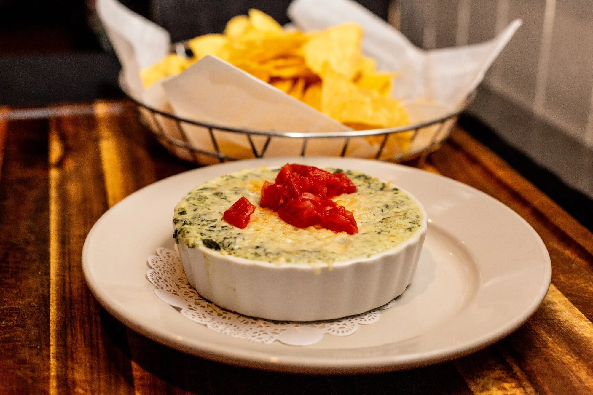 Spinach and cheese dip topped with chopped tomatoes served in a small white ramekin, with a basket of potato chips in the background.