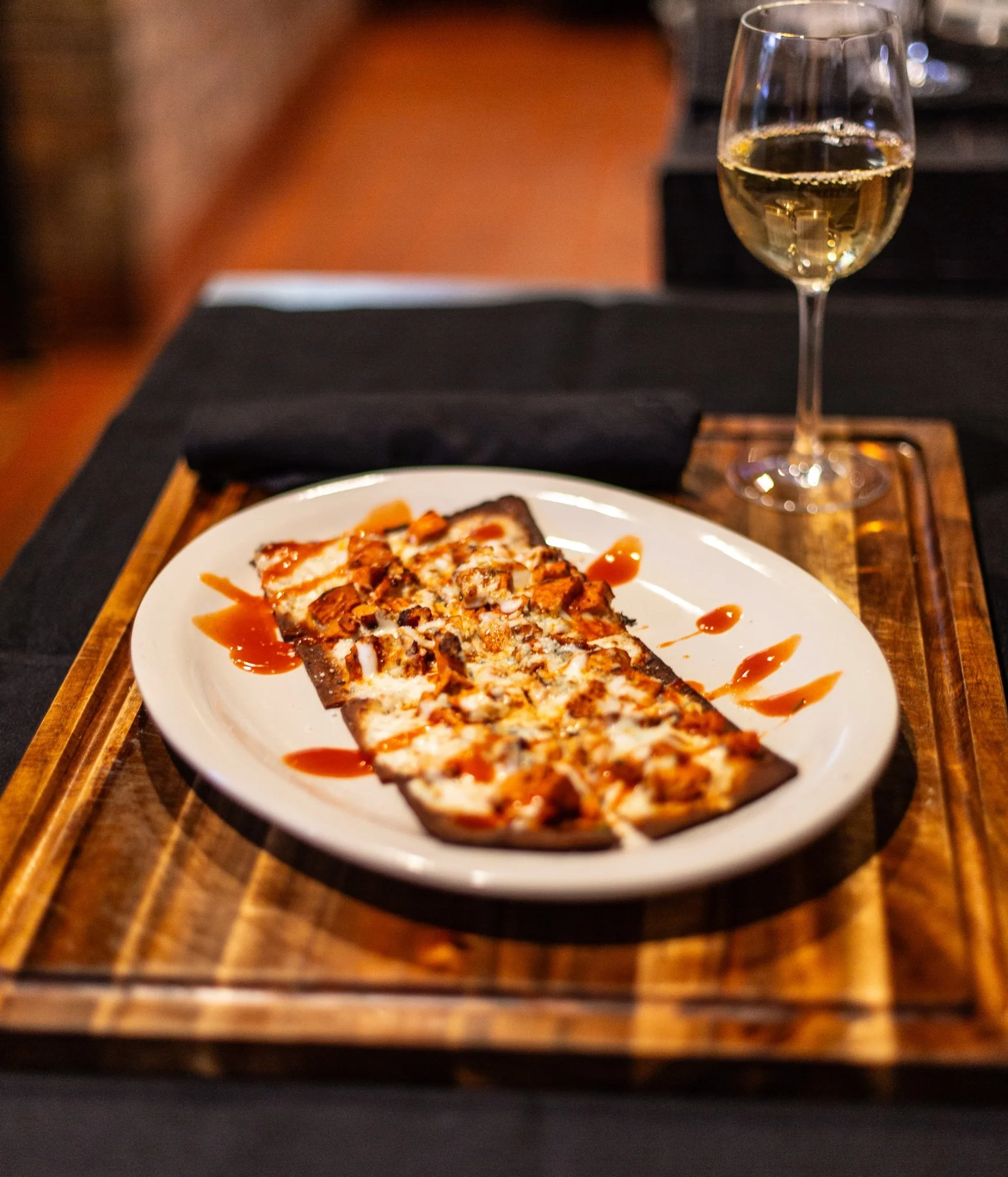 A flat bread pizza with cheese, toppings, and sauce on a white plate, served with a glass of white wine on a wooden tray.