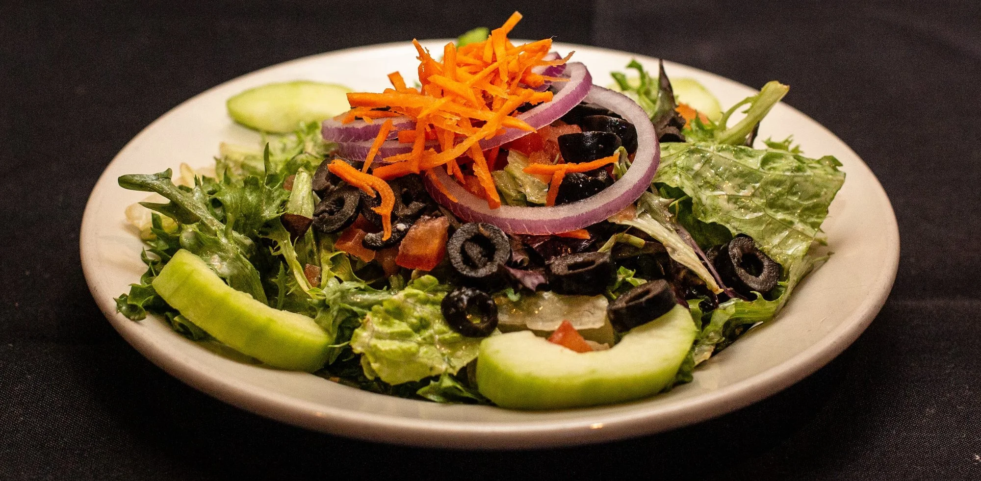 A fresh green salad with sliced cucumbers, shredded carrots, red onion rings, black olives, and various leafy greens on a white plate.