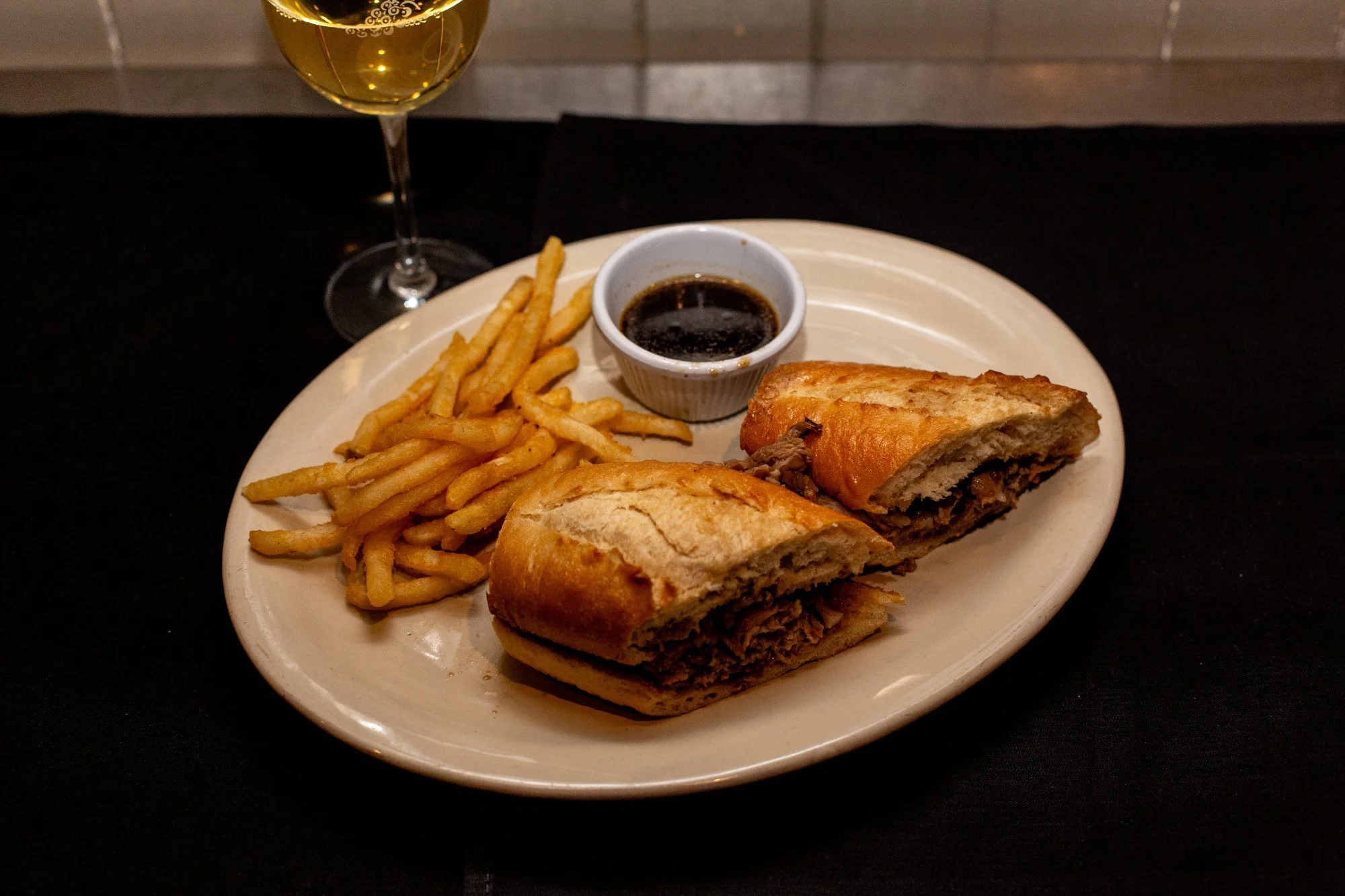 Plate with French fries, a beef-filled sub sandwich, a small bowl of dark dipping sauce, and a glass of white wine.