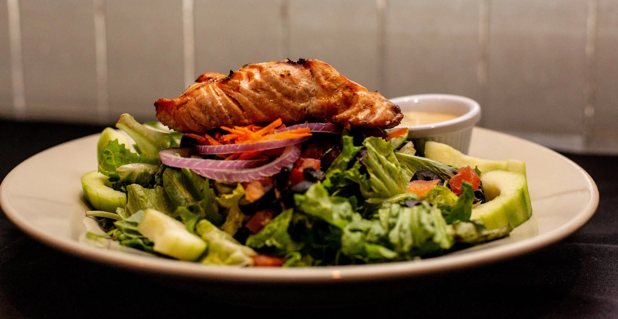 Grilled salmon served on a bed of mixed greens and sliced cucumbers, with a small container of dressing on the side.