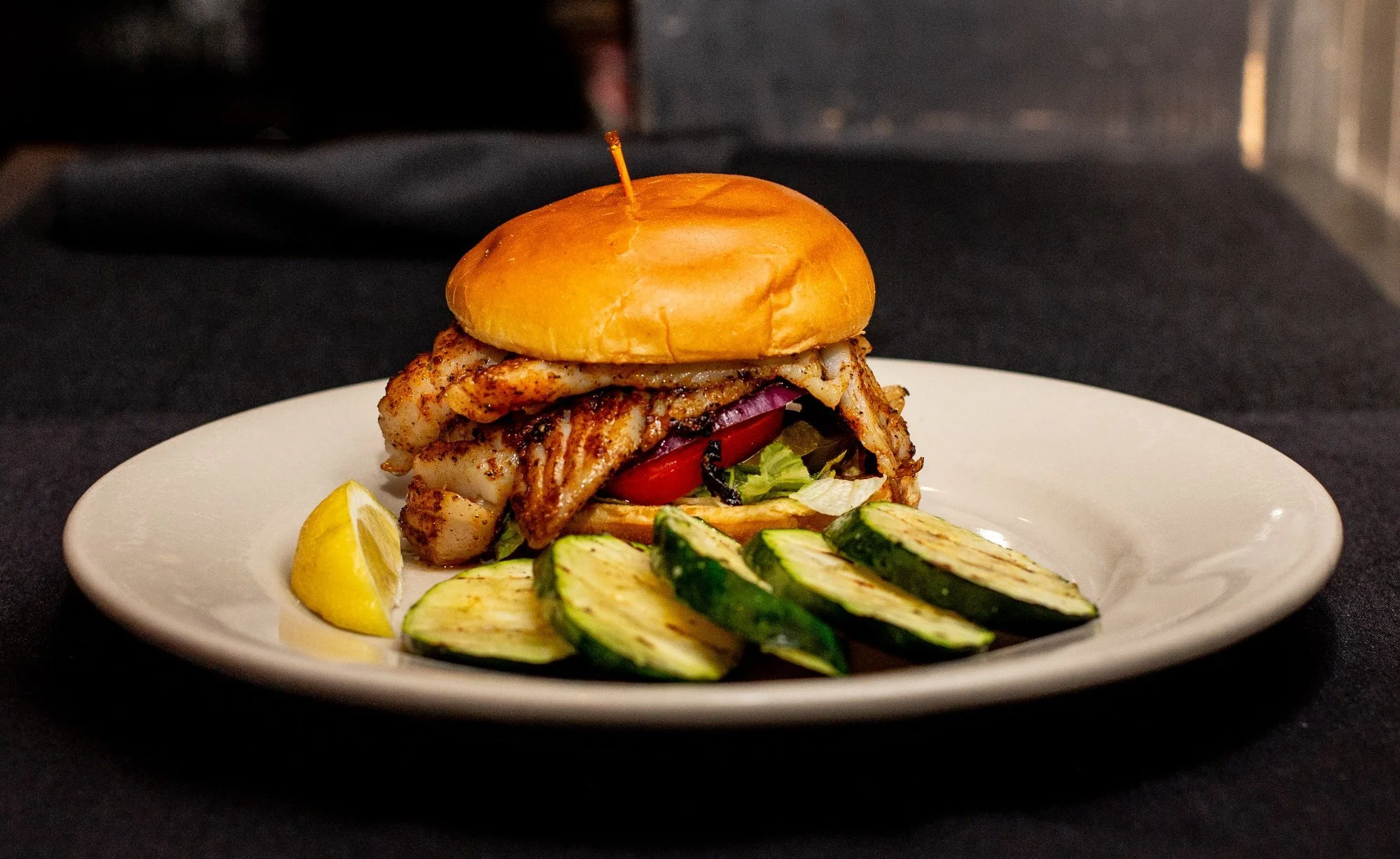 Grilled grouper fish sandwich with lettuce, tomato, onion, and a bun served with grilled zucchini slices and a lemon wedge on a white plate