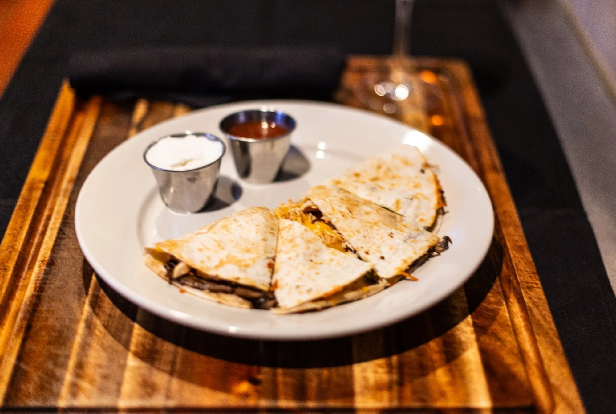 Quesadilla slices on a white plate with two small cups of sour cream and salsa, served on a wooden tray.