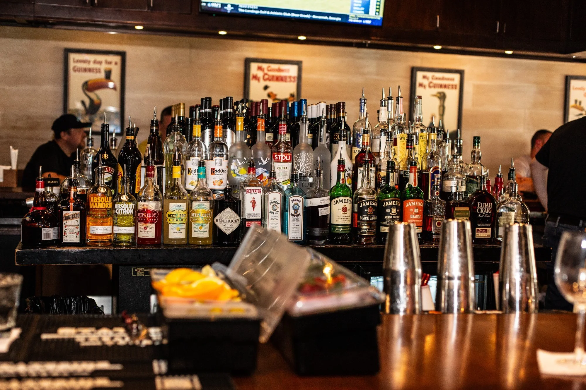 A bar with various bottles of alcohol on the countertop, including whiskey, gin, and vodka, with cocktail shakers and bar tools in the foreground.