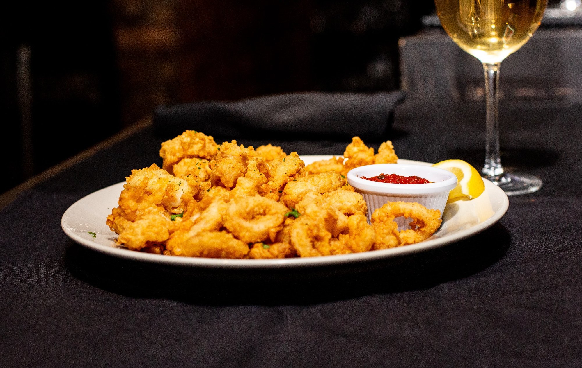 Plate of fried calamari with a lemon wedge, marinara sauce, and a glass of white wine on a black tablecloth.