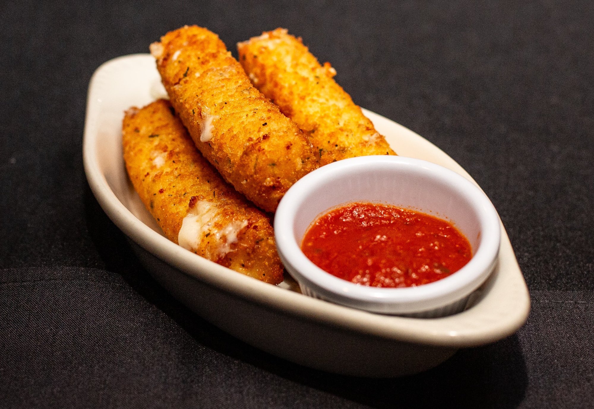 Three mozzarella sticks with marinara sauce in a small white dish, on a white oval plate against a black background.