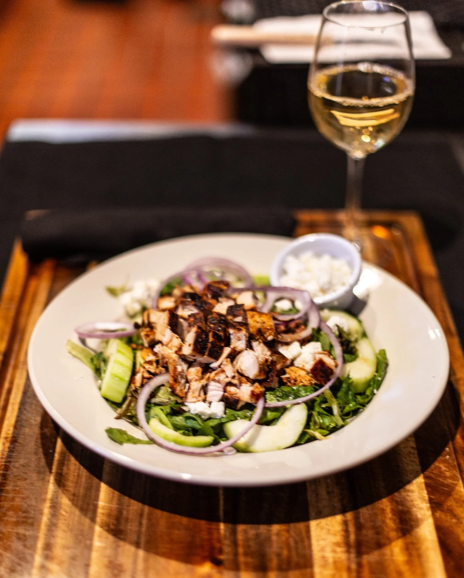 A Greek salad with grilled chicken, cucumbers, red onions, feta cheese, and herbs, served with a glass of white wine.
