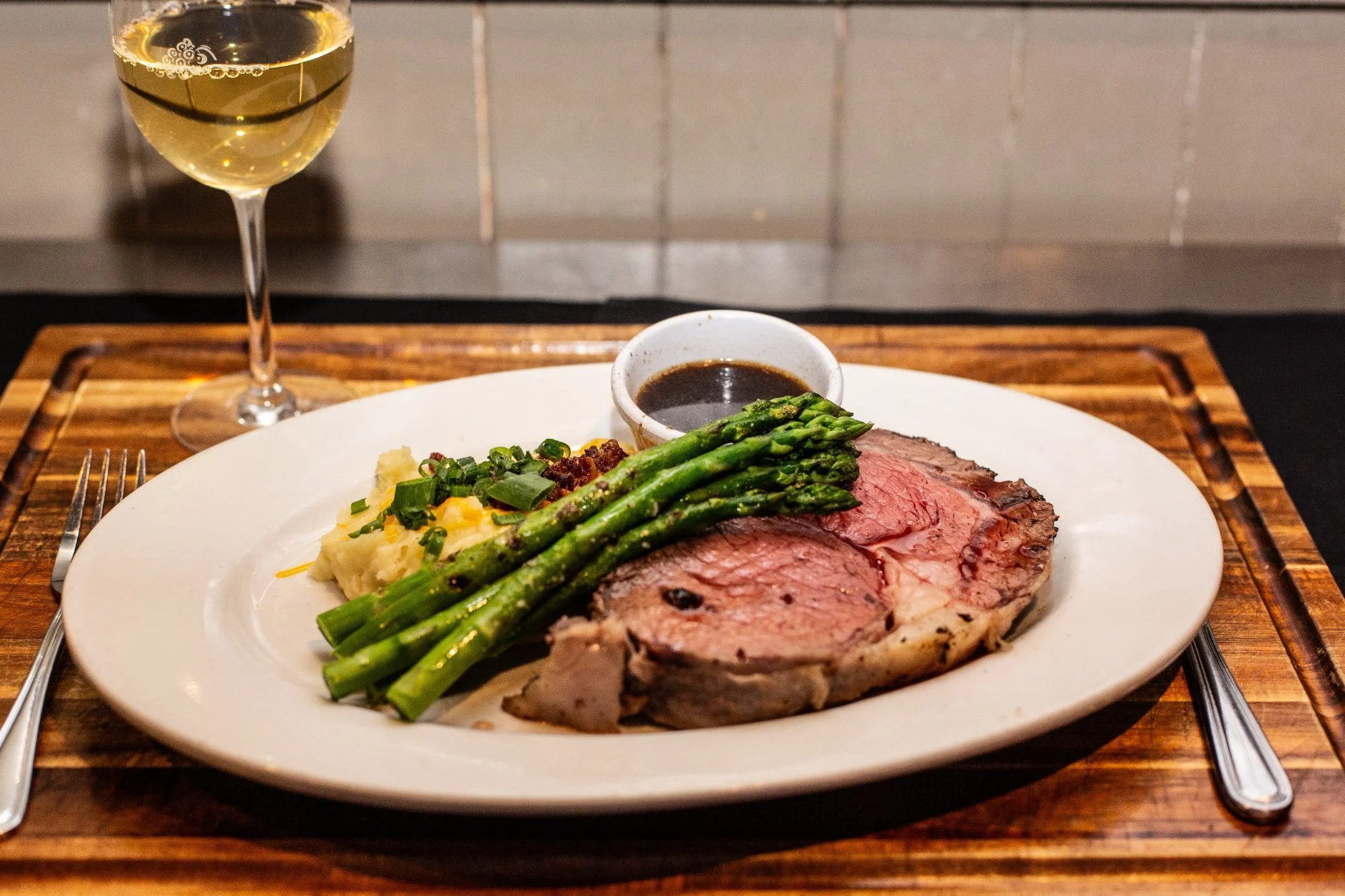 A dinner plate with slices of beef Wellington, green asparagus, mashed potatoes topped with green onions, and a small dish of sauce. A glass of white wine is in the background.