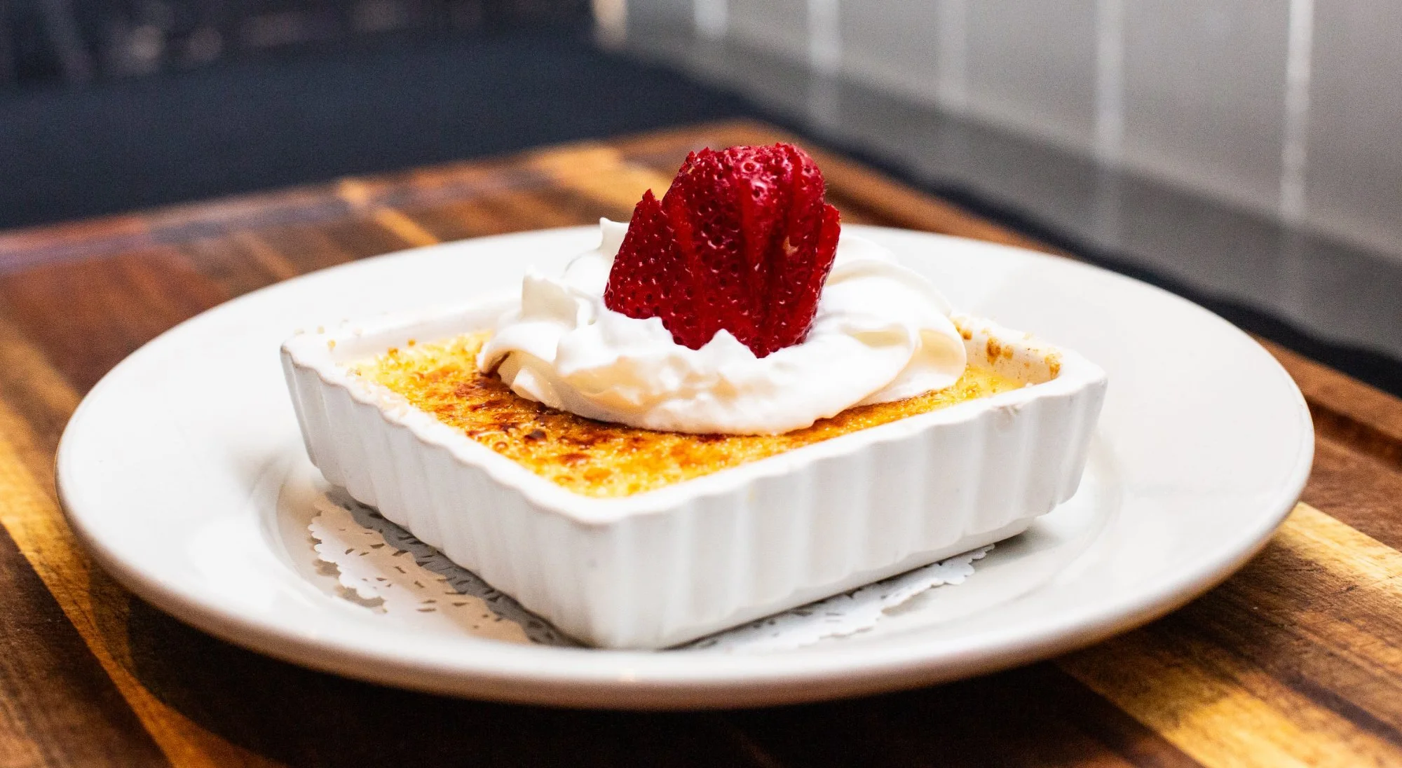 A serving of crème brûlée topped with whipped cream and a strawberry, presented in a white square ramekin on a white plate.