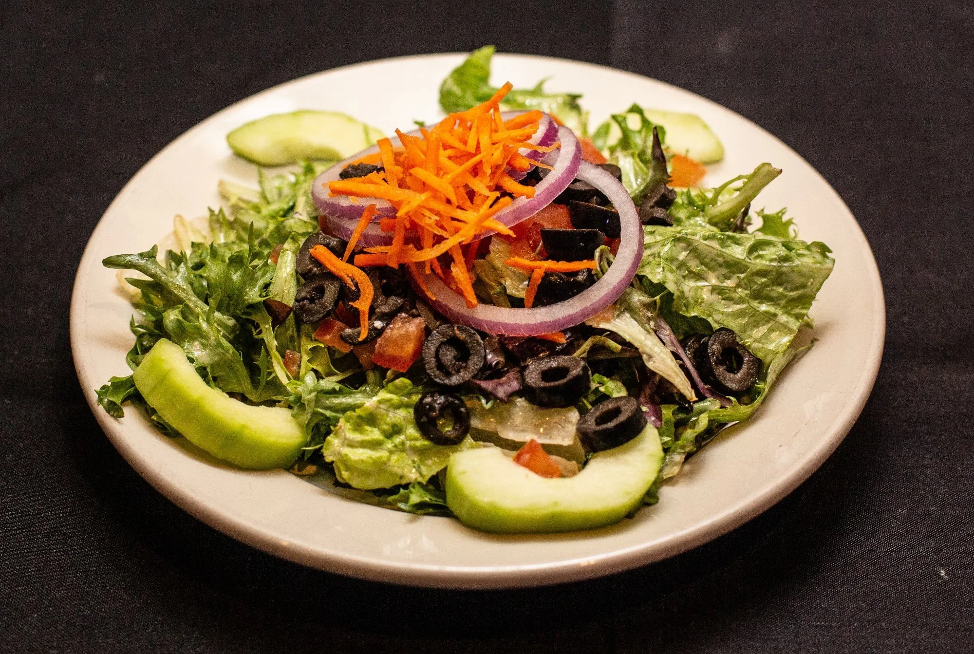 A fresh vegetable salad with lettuce, sliced cucumbers, sliced red onions, black olives, shredded carrots, and diced tomatoes on a white plate.
