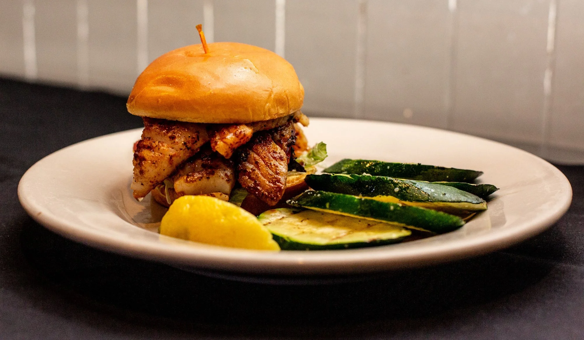 A plate with a sandwich topped with grilled grouper, along with grilled zucchini slices, and a lemon wedge on a black surface.