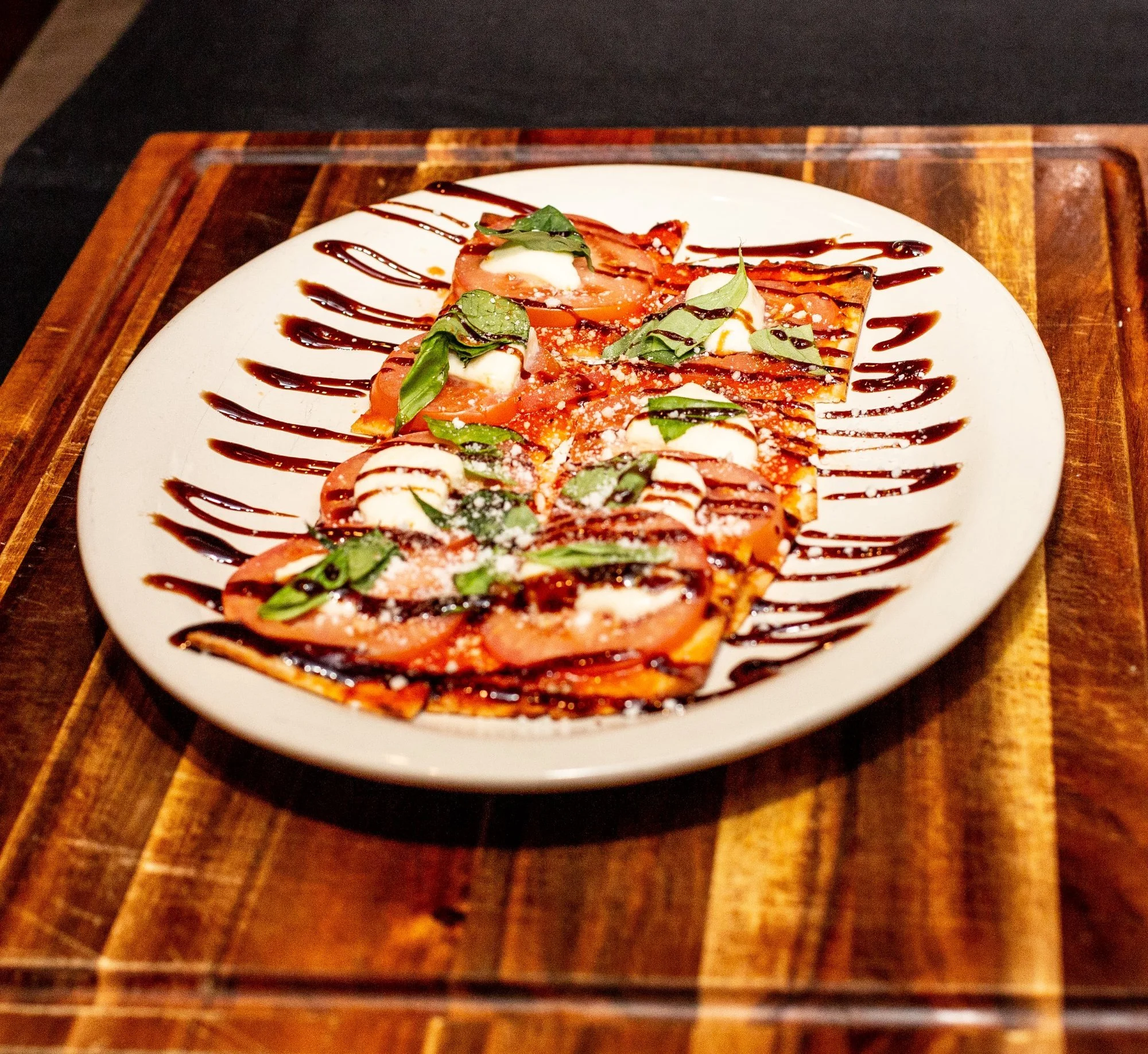 A plate of sliced tomatoes with mozzarella cheese, basil leaves, grated cheese, and drizzled with balsamic glaze and olive oil. The dish is served on a white oval plate on a wooden tray.