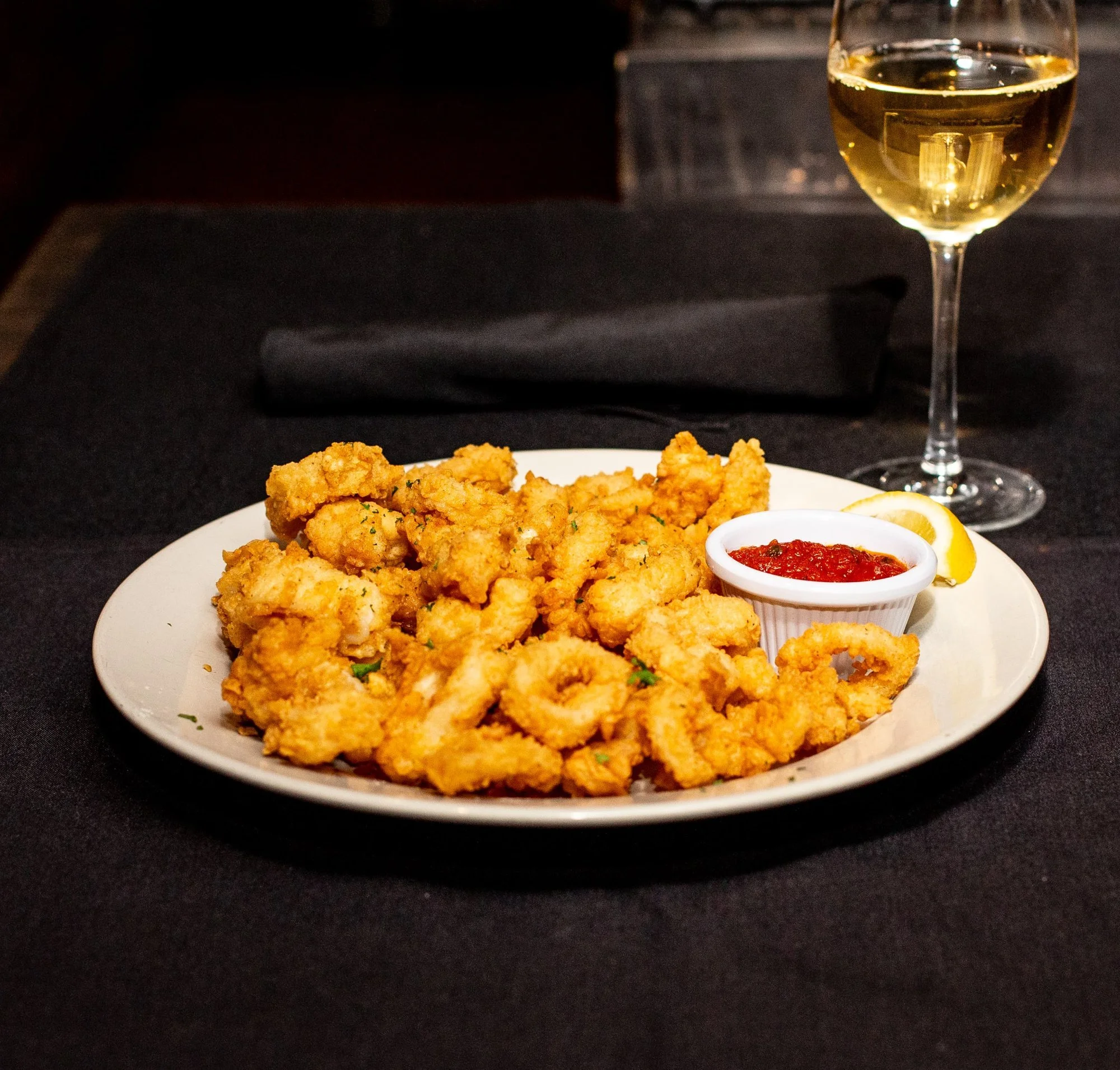 A white plate of fried calamari with a small bowl of marinara sauce, lemon wedges, and a glass of white wine on a black tablecloth.