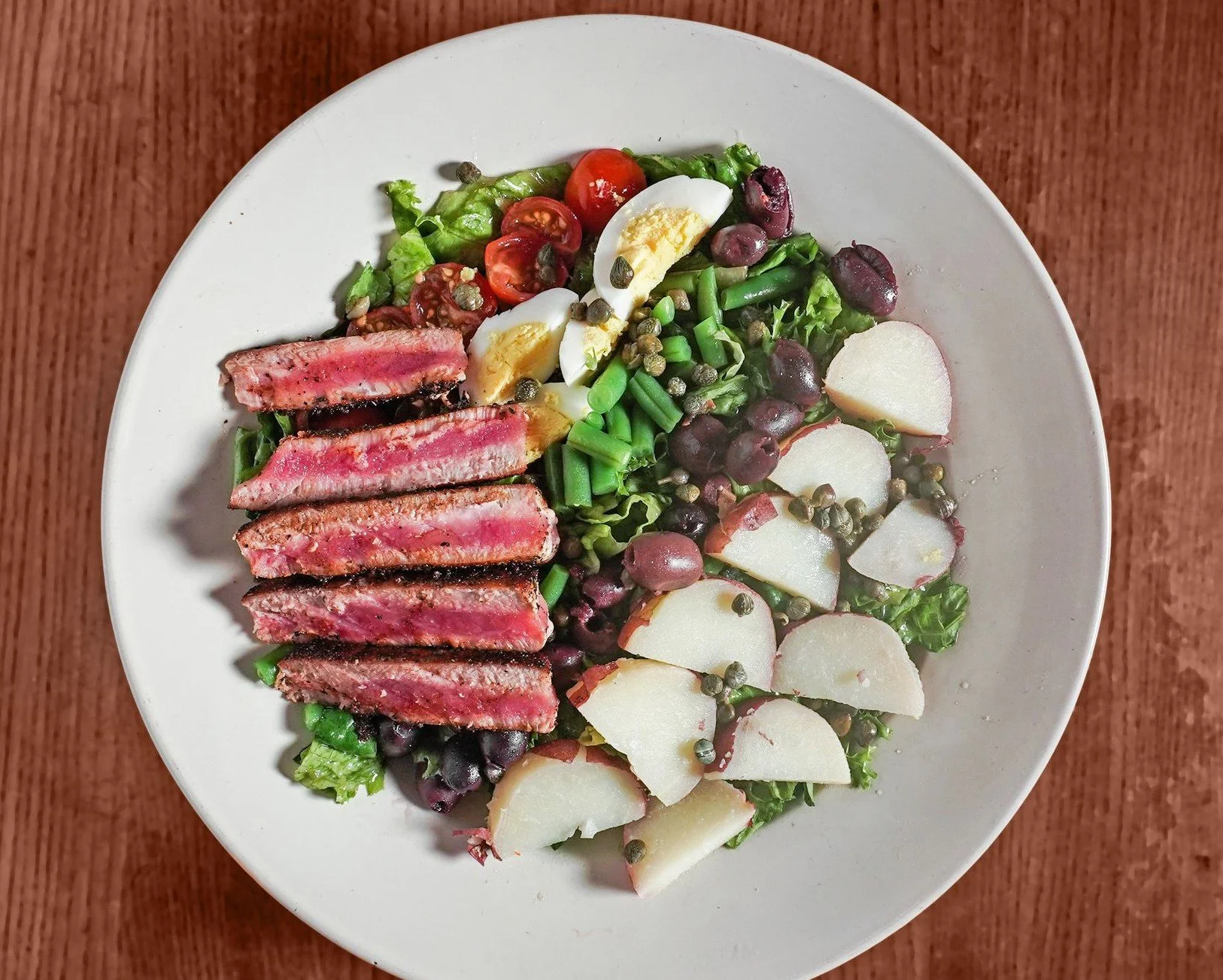 A white plate of mixed greens topped with cherry tomatoes, sliced boiled eggs, radish slices, grapes, green beans, and capers, on a wooden surface.