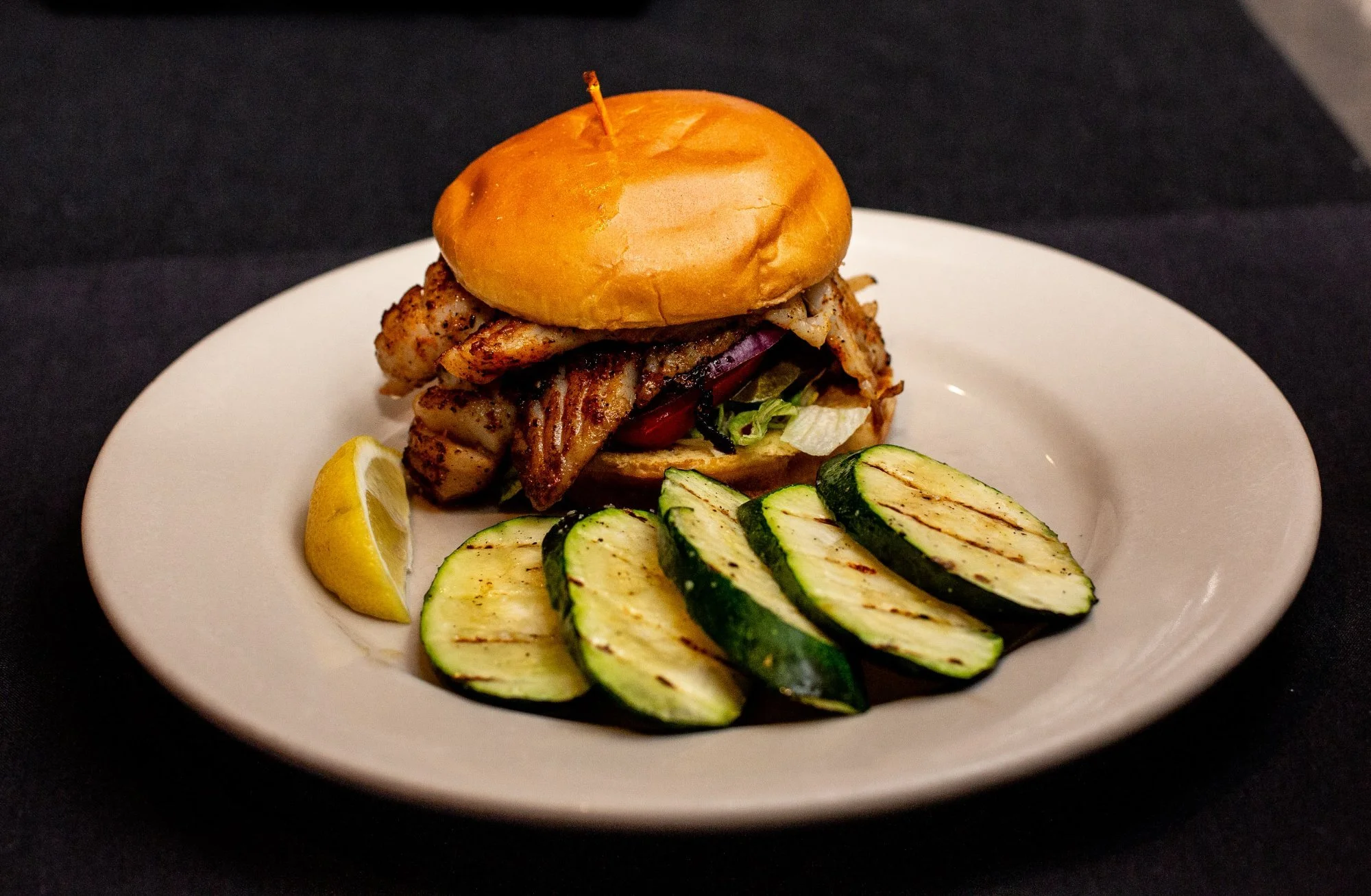 Grilled Grouper fish sandwich with lettuce, tomato, and grilled zucchini slices on a white plate, garnished with lemon wedge, on a black table.