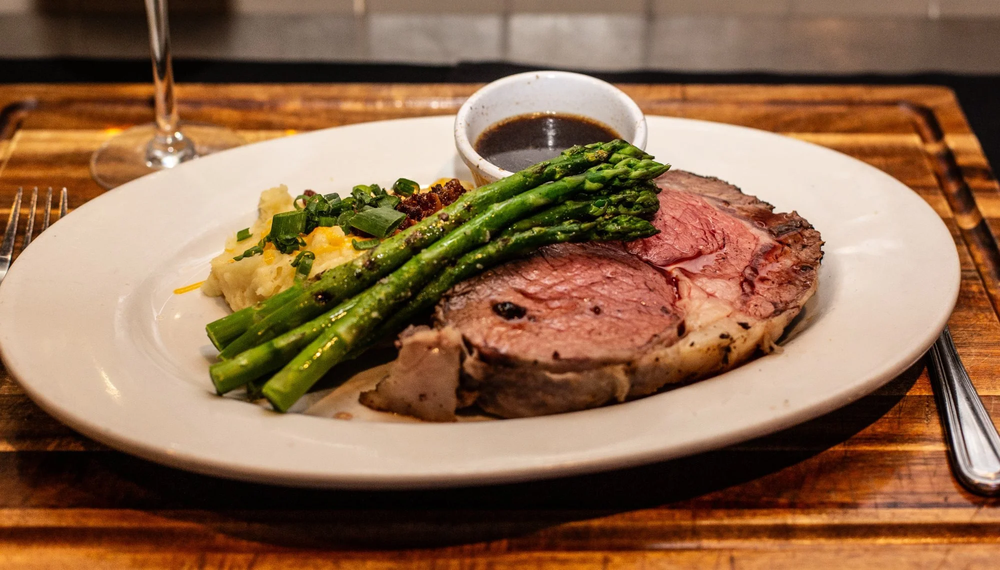 A plate of cooked beef steak, green asparagus, mashed potatoes with chives, and a small bowl of dark gravy, set on a wooden table with a wine glass and utensils.