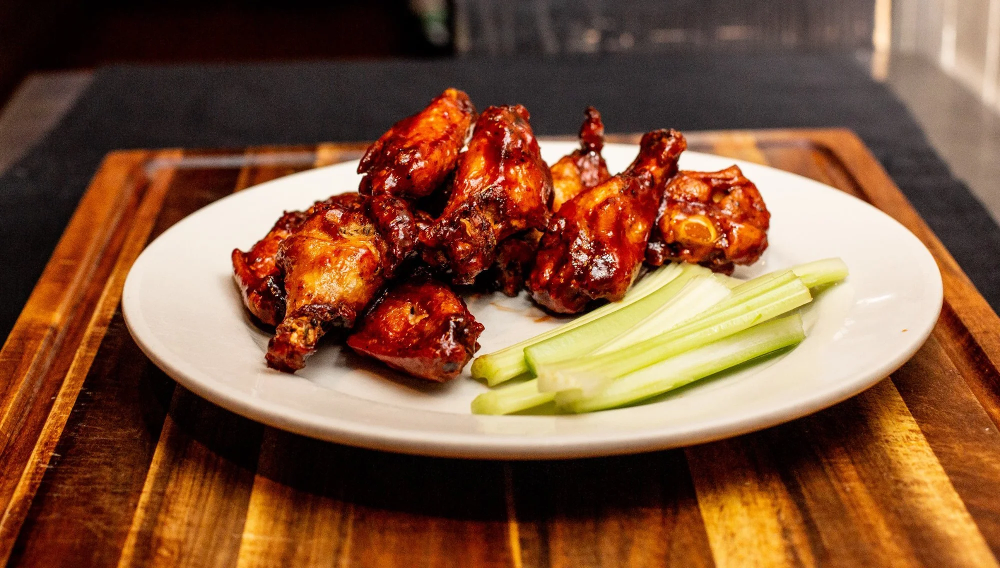 A white plate with glazed chicken wings and celery sticks on a wooden surface.