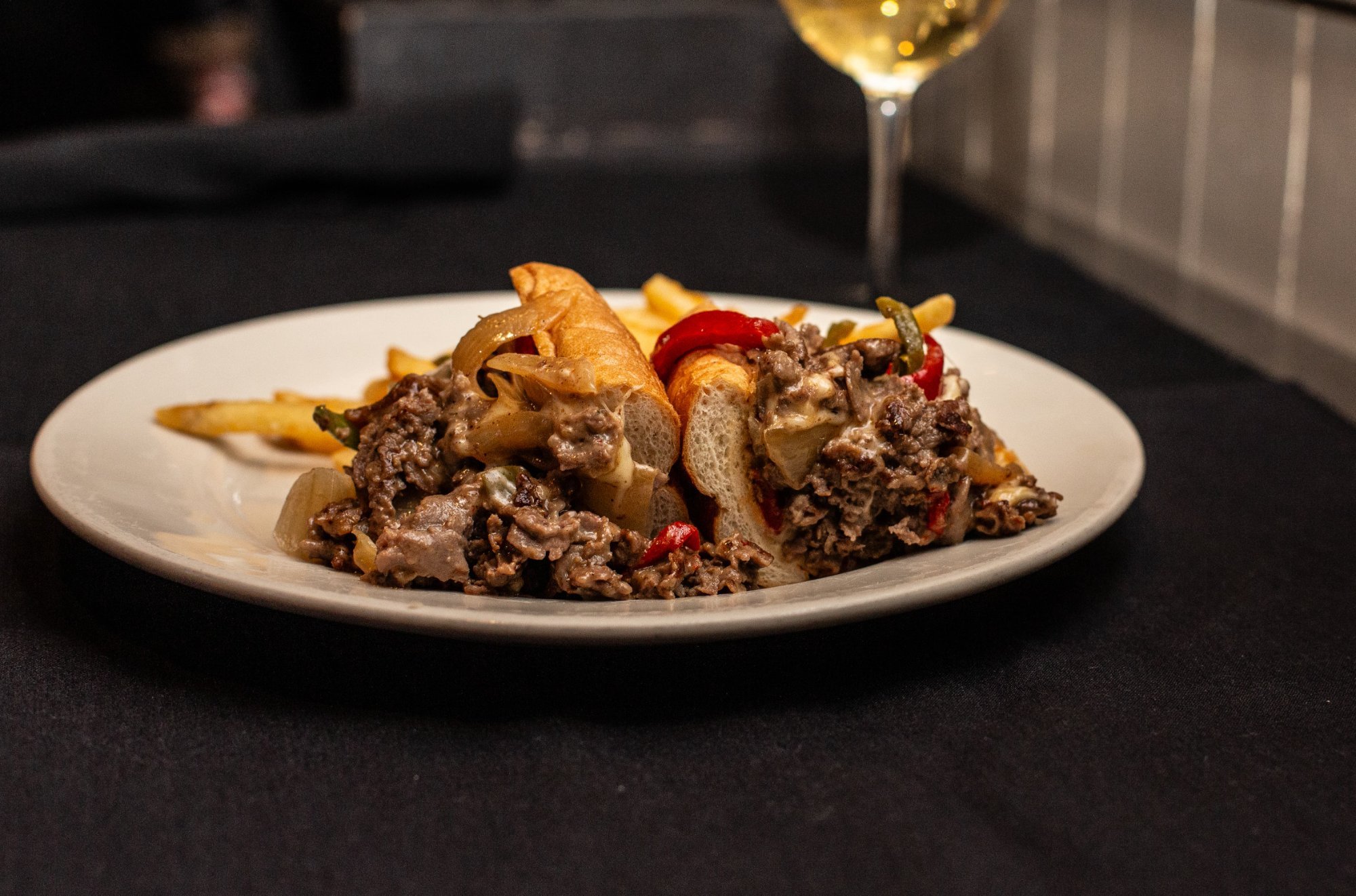 Plate with a Philly Cheesesteak sandwich, served with French fries and a glass of white wine in the background.