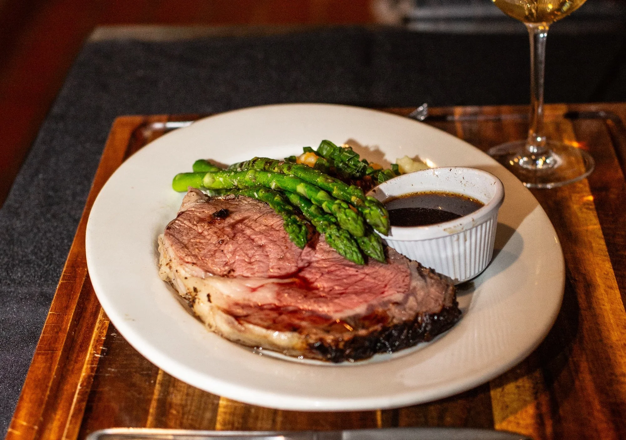A plate with a serving of medium-rare steak, garnished with grilled asparagus and green onions, alongside a small bowl of dark sauce. A glass of white wine is partially visible in the background.